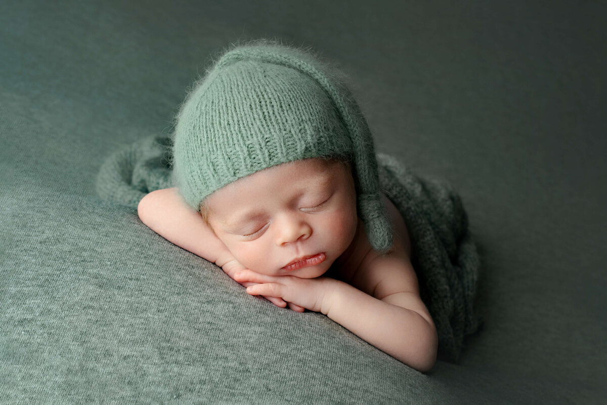 newborn boy laying with head on arms with  green  sleepy cap on green backdrop