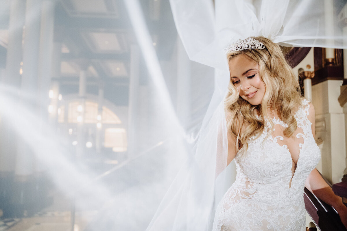 A bride in a lace wedding gown smiles as her veil flows gracefully in a beautifully lit, elegant setting, capturing the essence of luxury wedding design.