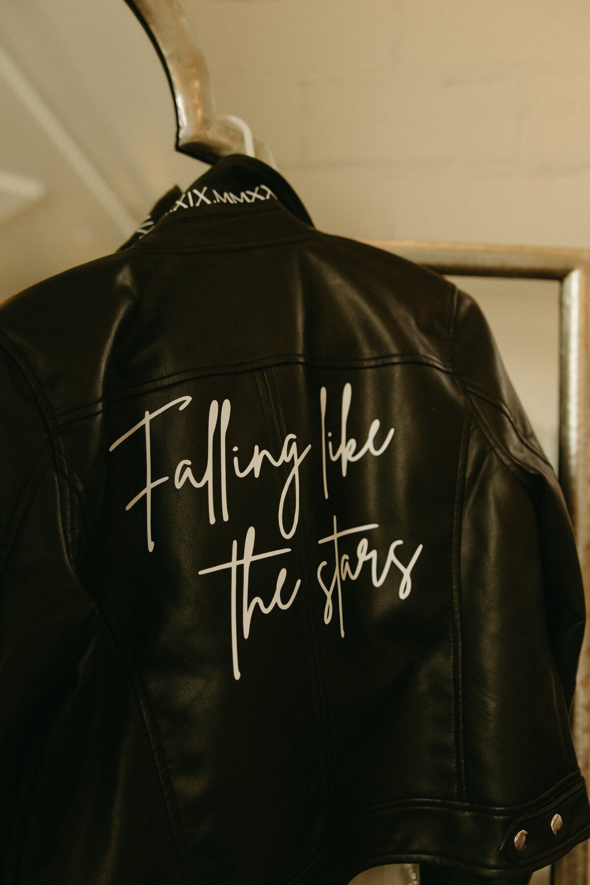 A black leather jacket hanging on a rack with the phrase "falling like the stars" written in white cursive on the back, perfect for park farm winery weddings.