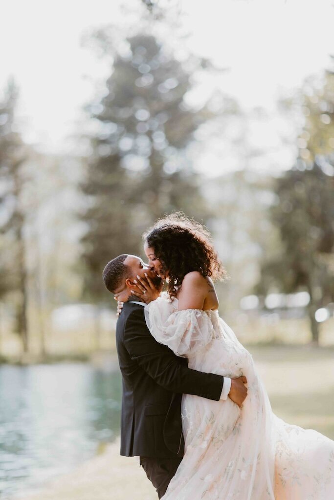 A groom lifts and kisses his bride at sunset by a lake