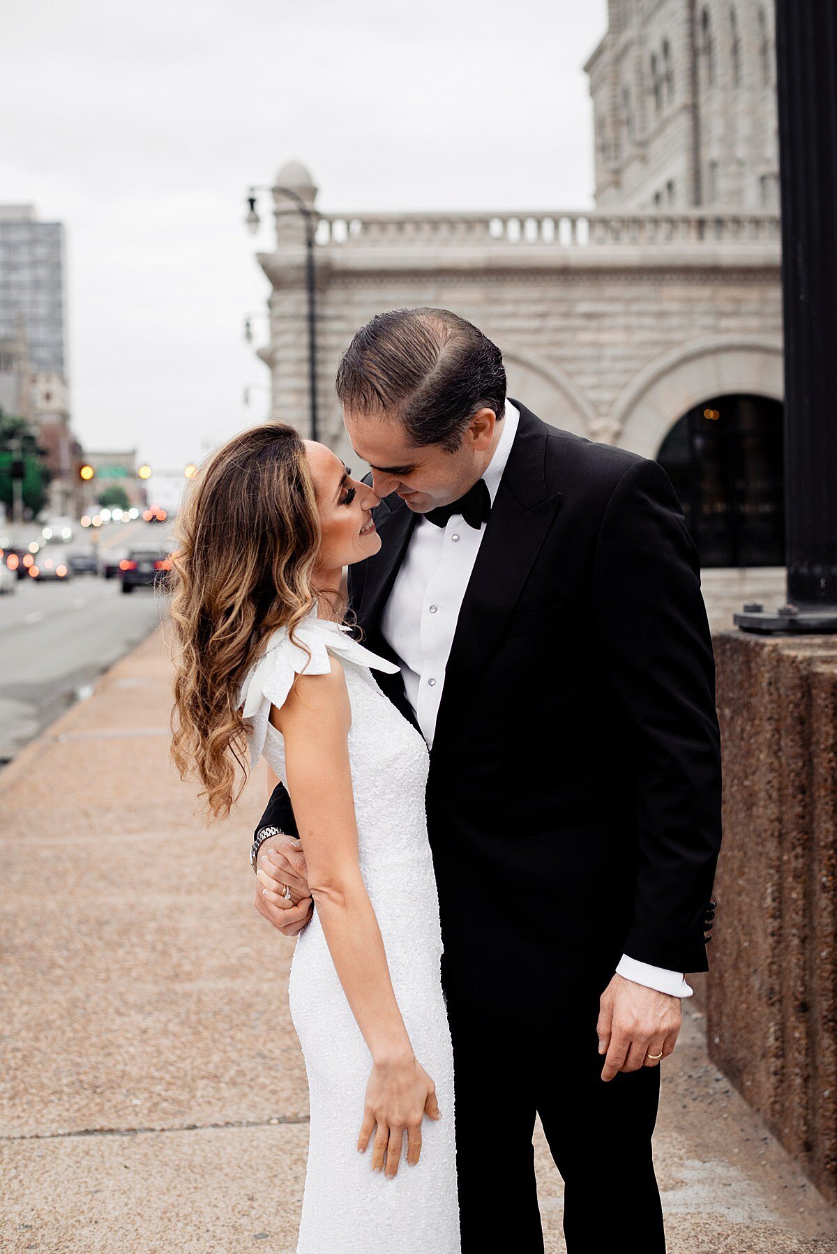 The groom leans in for a kiss as he wraps one arm around the bride's waist. They are standing outside of historic Union Station in Nashville. The bride is wearing a one shoulder sheath dress with a ruffle over her shoulder. The groom is wearing a black tuxedo with a white shirt and black bowtie.