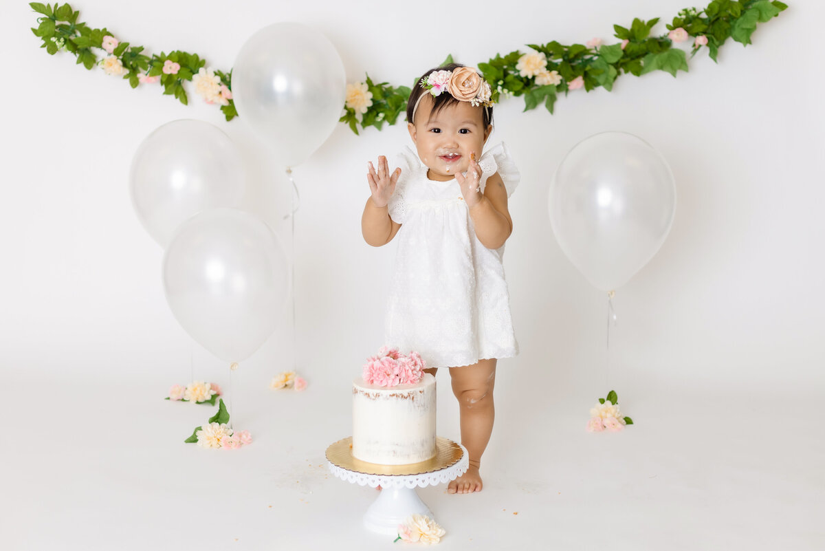 Cake Smash Photographer, a baby girl stands before a cake and balloons