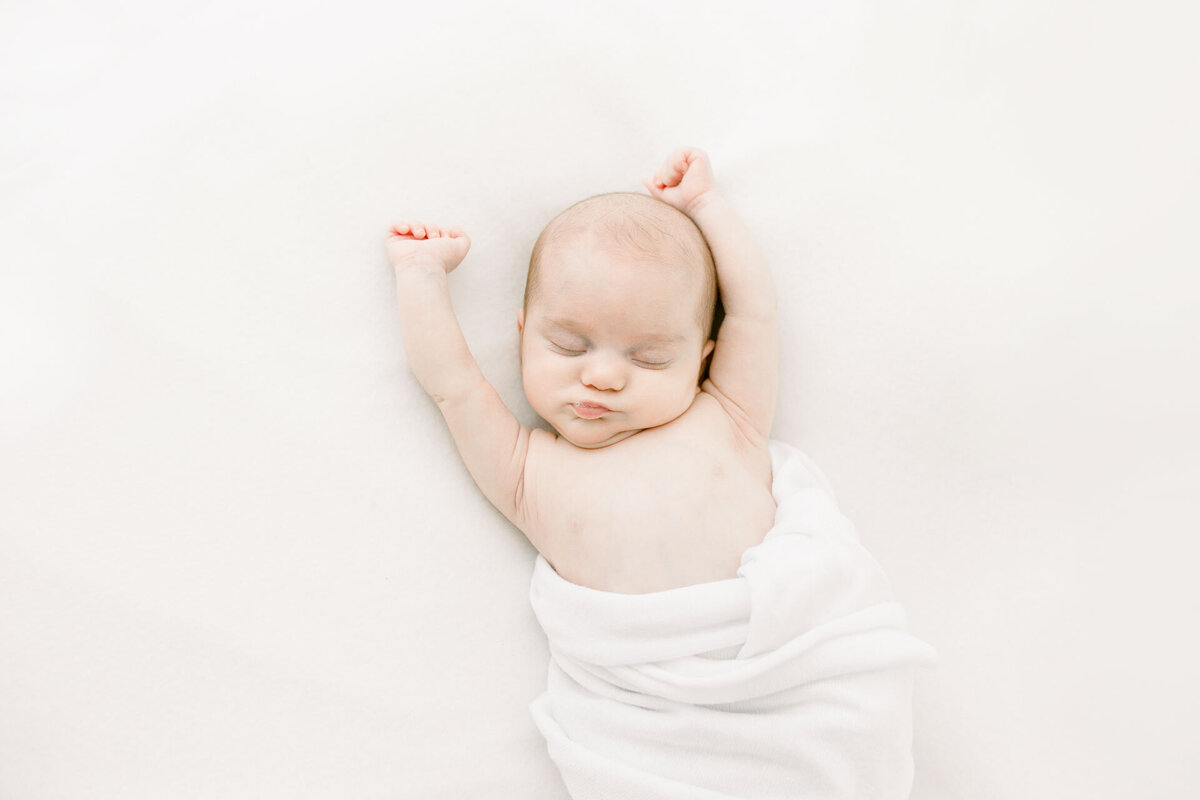 Babyshooting: Neugeborenes schlafend mit ausgestreckten Armen in weißer Decke auf weißem Untergrund.