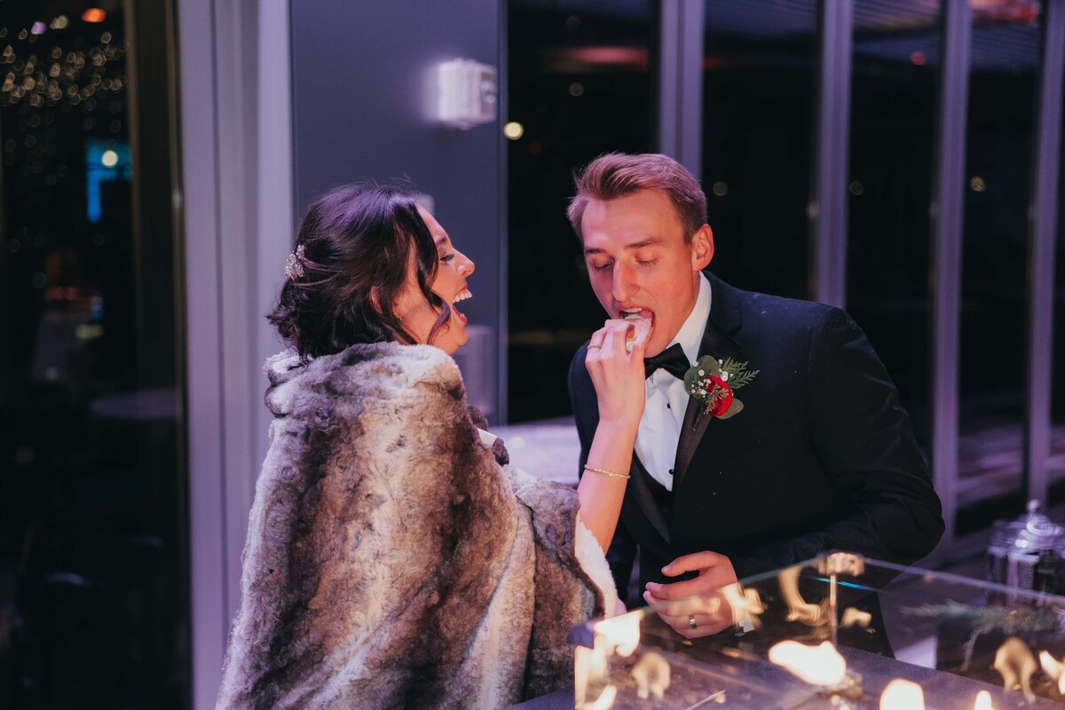 A newlywed couple in elegant attire share a romantic moment by a candlelit table at a winery, with the groom feeding the bride a bite of dessert.