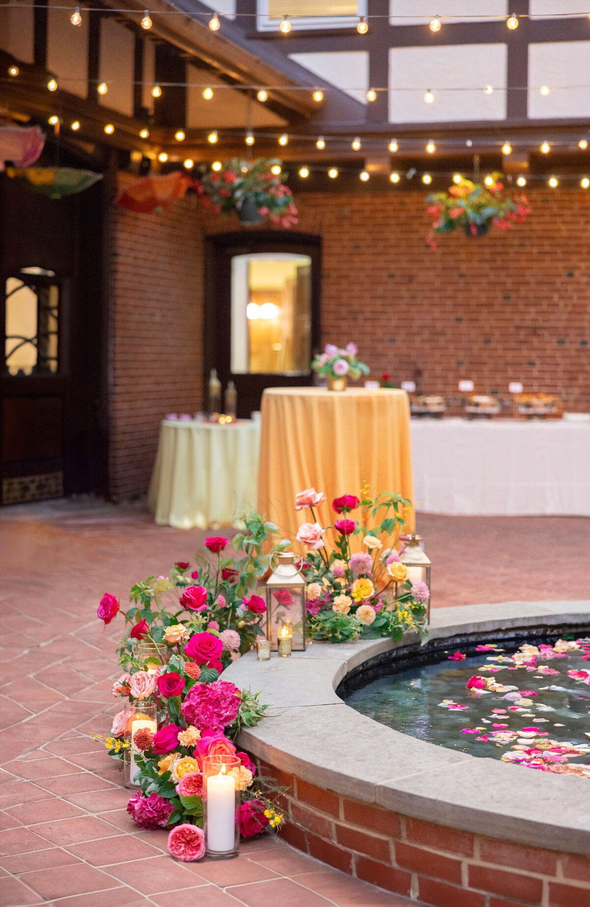 A courtyard set up for an event, featuring a brick circular fountain with floating petals, surrounded by lanterns and colorful flowers. Tables with tablecloths and food are in the background, under string lights.