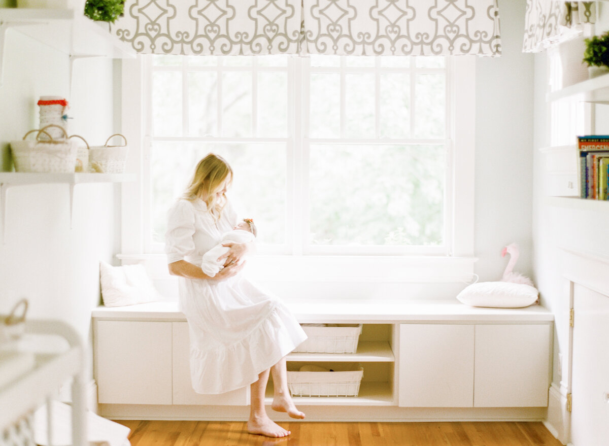 Mom sits in a window seat while holding her newborn. Photographed by Raleigh Newborn Photographer A.J. Dunlap Photography.