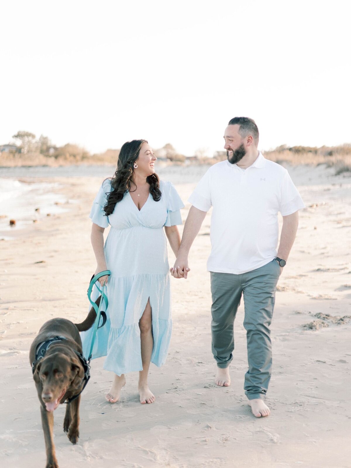 Man and woman walk along beach with dog smiling at each other.