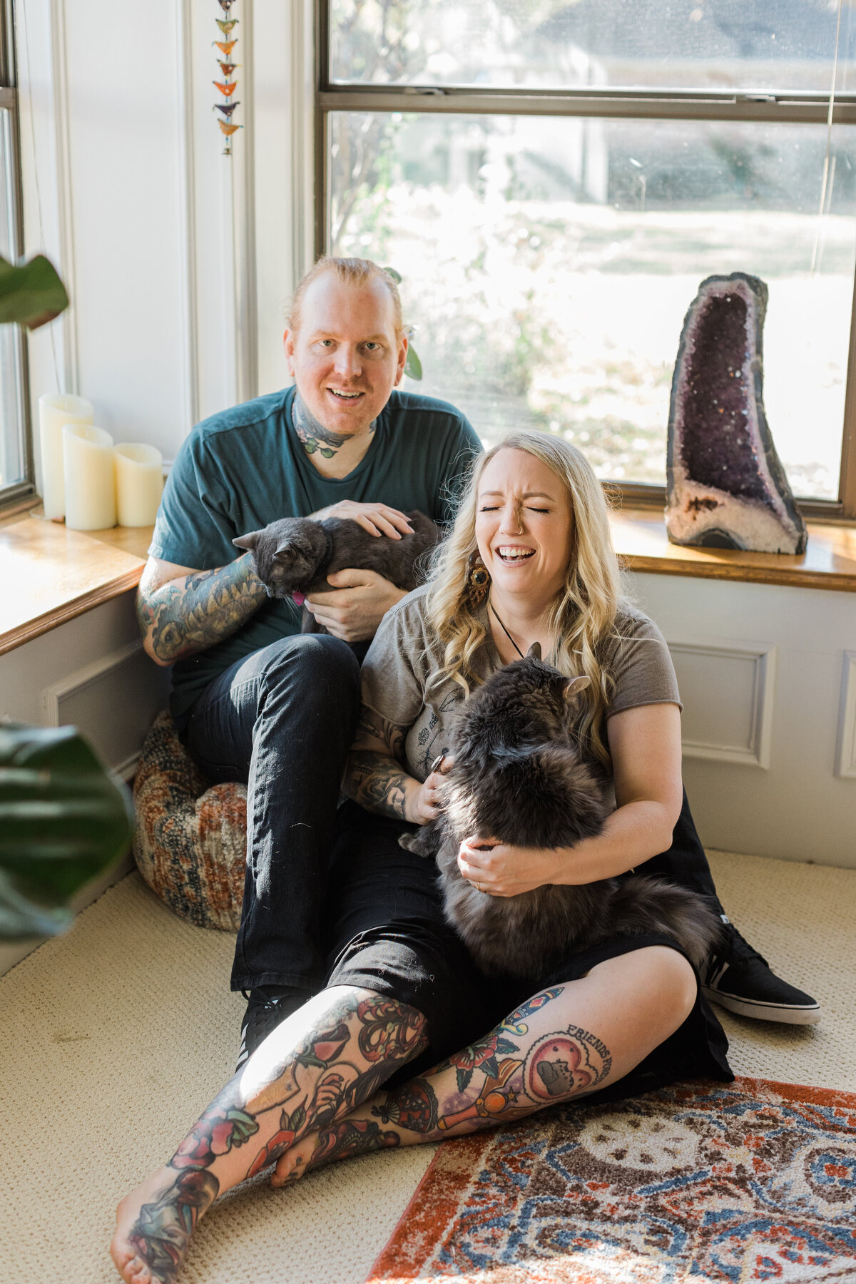 A couple casually posing with their two cats in their home during their engagement session in DFW, Texas. The woman on the right is seated on the floor and is laughing with a large, fluffy cat in her lap. She is wearing a t-shirt and shorts and has many tattoos on her legs and right arm. The man on the left is seated on a cushion and is holding and petting a short hair cat. He's wearing a t-shirt and pants and is also covered in many tattoos. A large geode sits beside them on a windowsill.