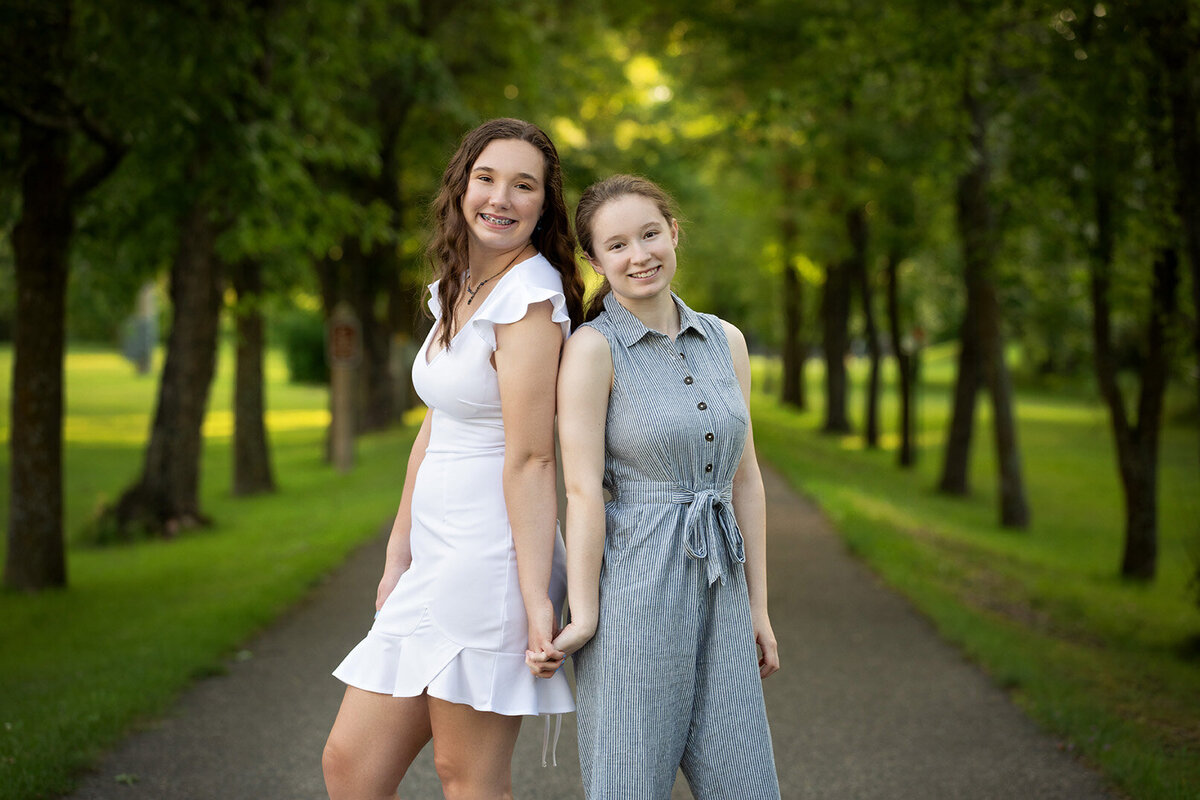 Fraternal twin sisters standing back to back holding hands on bike path in Akeley Minnesota during summer time.