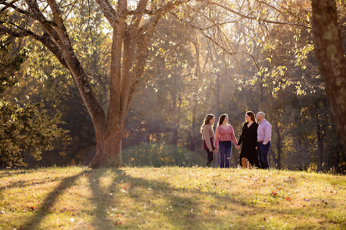 Radiant autumn family portrait on grassy hill in Fort Mill SC, parents and two daughters smiling in golden evening light at Anne Springs Close Greenway