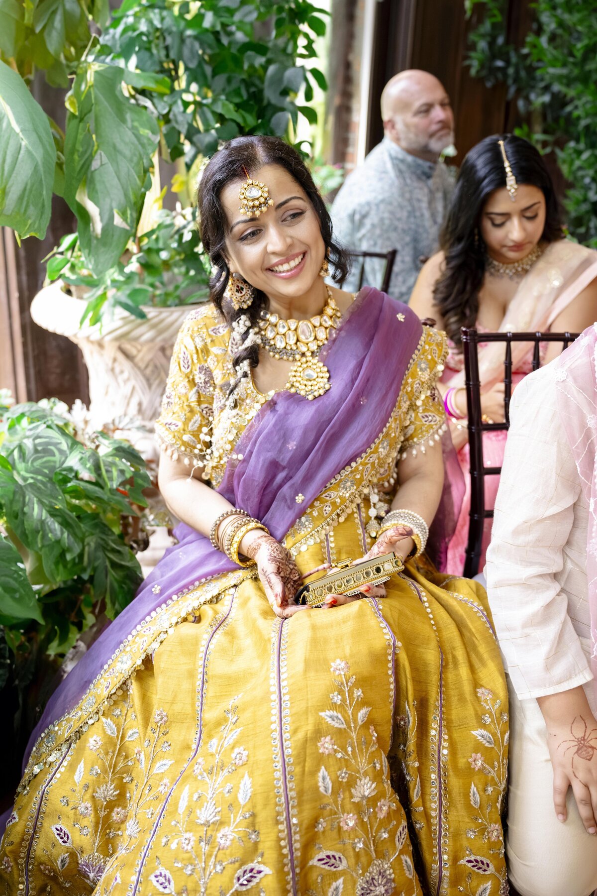 A woman in a yellow and purple traditional outfit sits and smiles. She is adorned with gold jewelry and surrounded by lush green plants. Another person sits nearby, partially visible. The setting appears festive and lively.