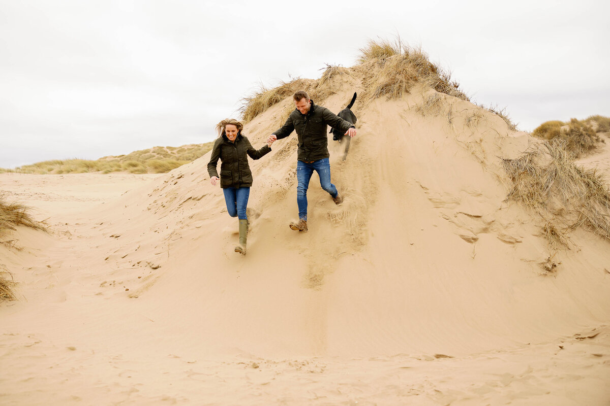 A pre wedding photograph of a couple and their dog running down a sand dune on Formby beach