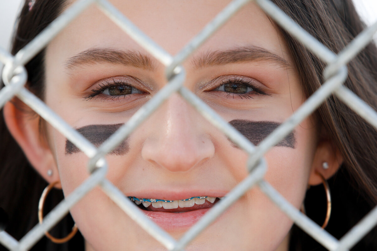 white teenage girl wears under eye black and looks through the metal fence at a softball field New York senior portrait photographer Milestone Images