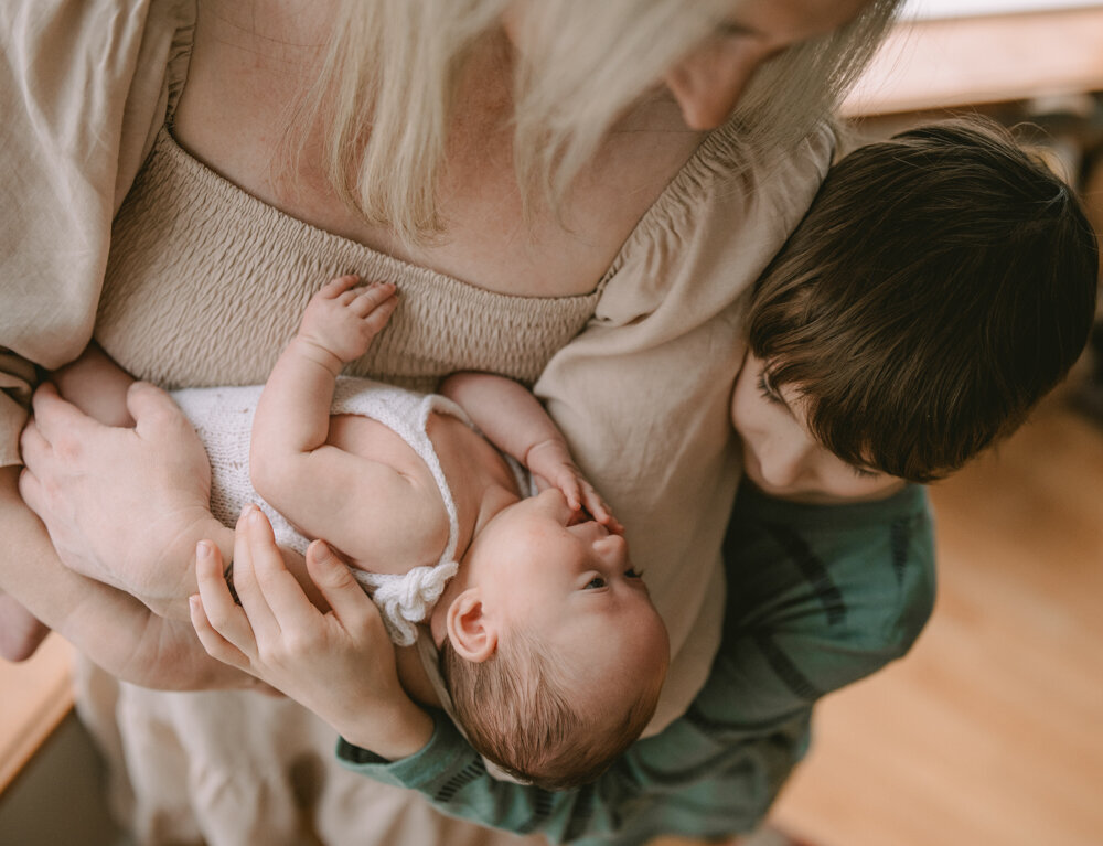 Big sibling and mother gazing at newborn in arms, captured in a soft and natural family moment during an in-home newborn session in Portland.