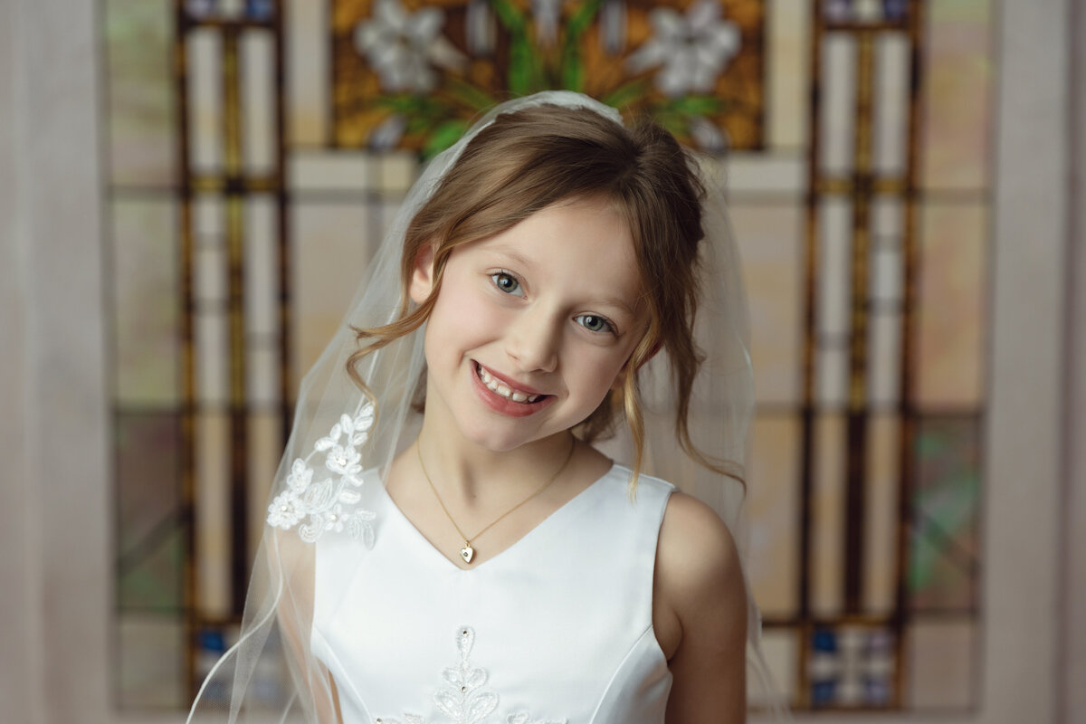 A young girl in a white communion dress stands by some stained glass smiling