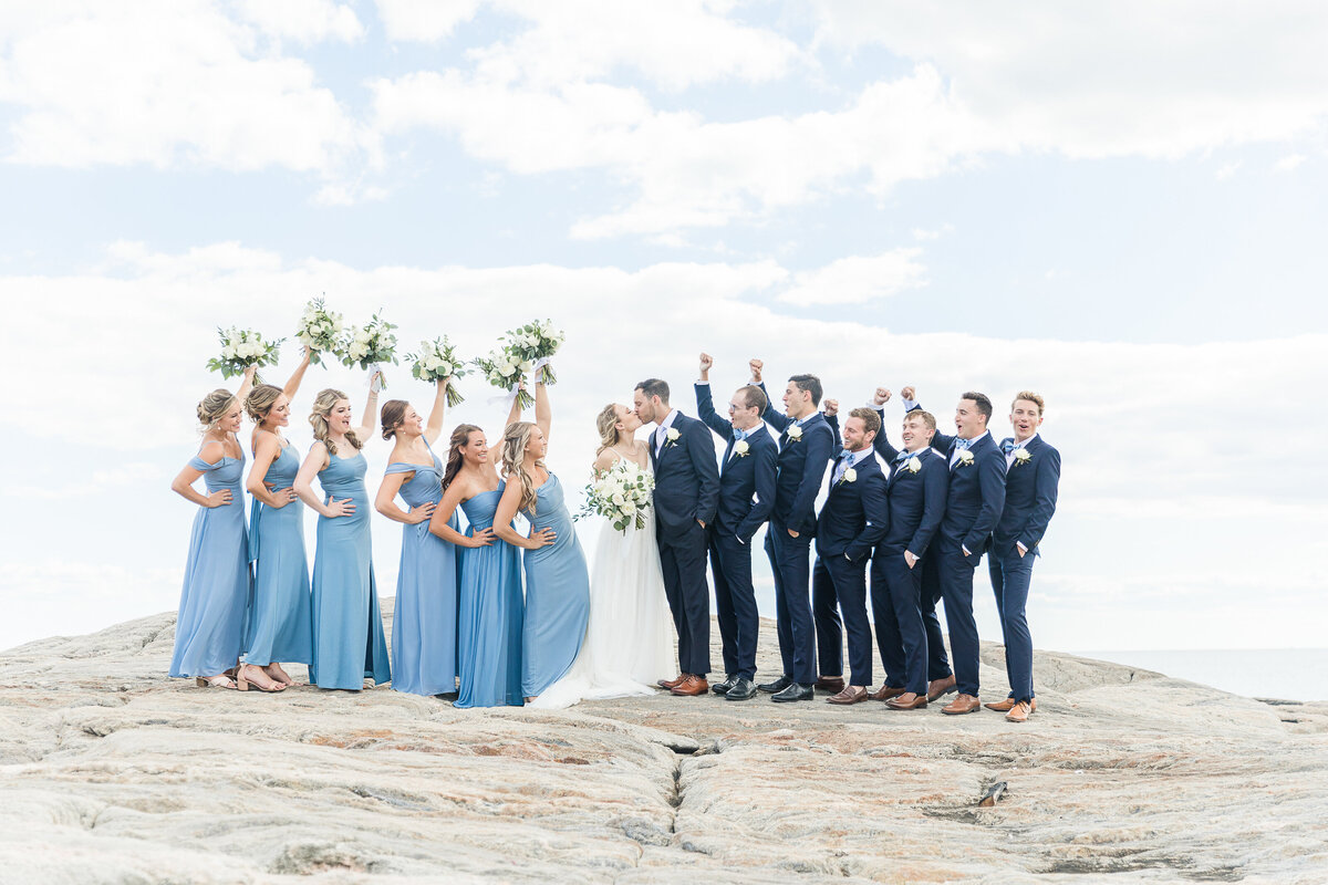 Bride and groom share a kiss on the rocks along the ocean shoreline at the Madison Beach Hotel. The bridesmaids and groomsmen have their hands raised in celebration. Captured by best New England wedding photographer Lia Rose Weddings.