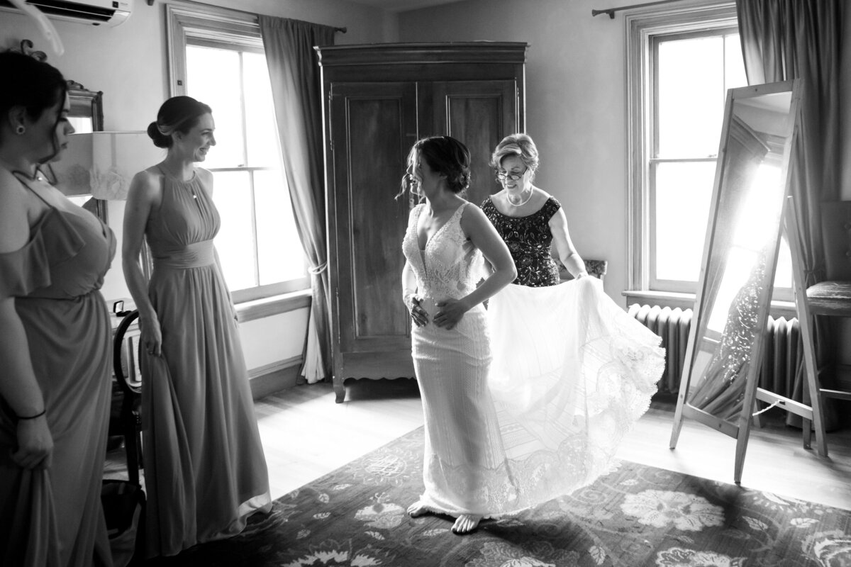 black and white photo of mother of bride helping daughter dress with two bridesmaids looking on Hudson Balley wedding photographer Milestone Images