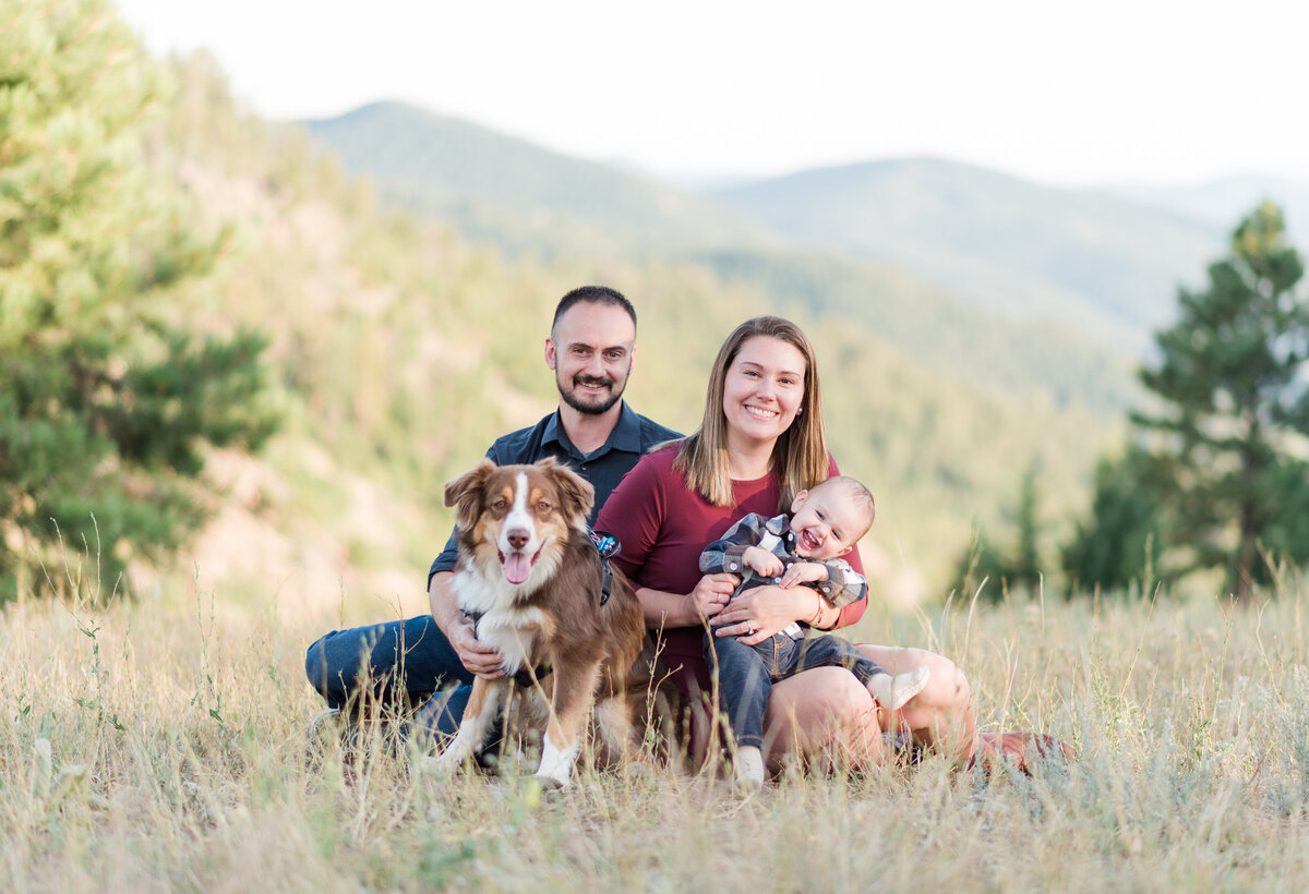 family photos denver with parents sitting a field with their baby and dog with the Rocky Mountains in the distance photographed by denver family photographers