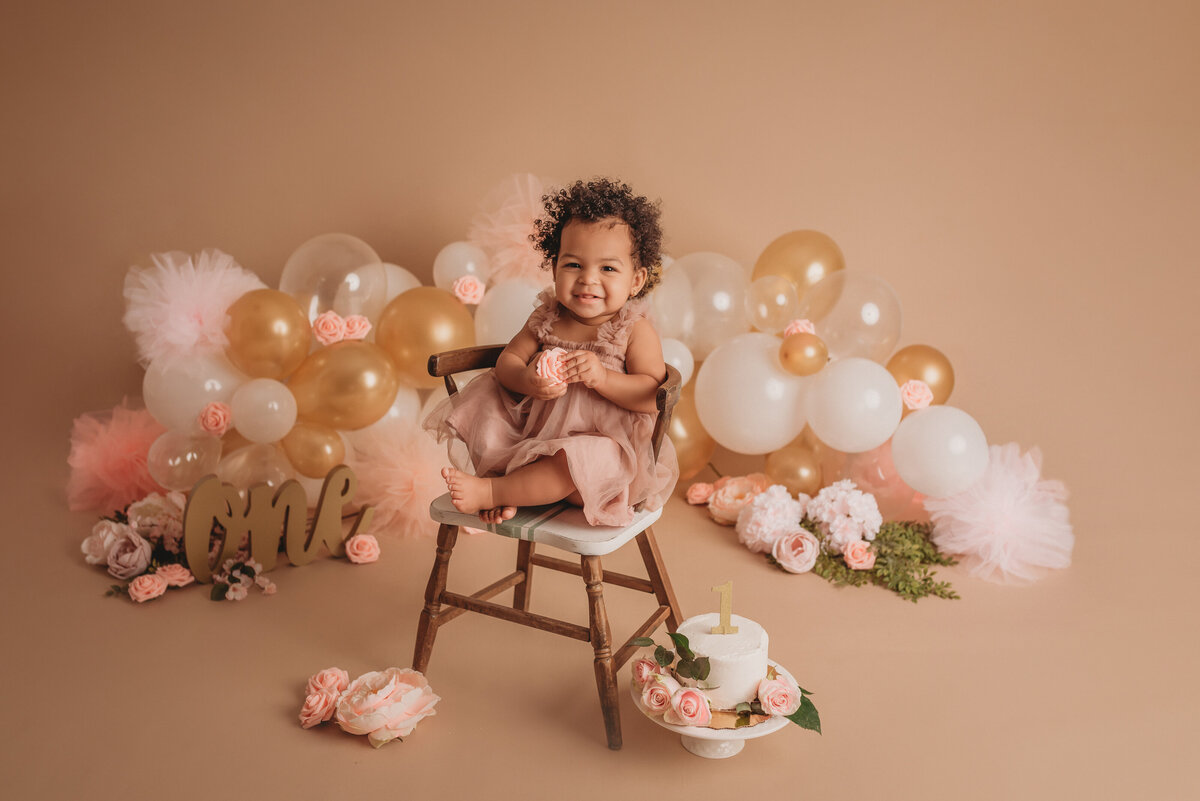 One year old baby girl wearing pink tulle dress sitting in miniature kids wooden chair smiling with birthday cake beside her and white, pink and gold balloon garland behind her on a tan backdrop
