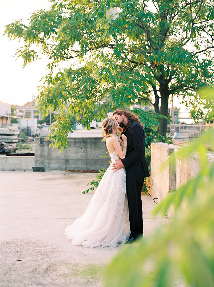 Bride and groom wearing a black tuxedo and white wedding gown kiss on an industrial rooftop.
