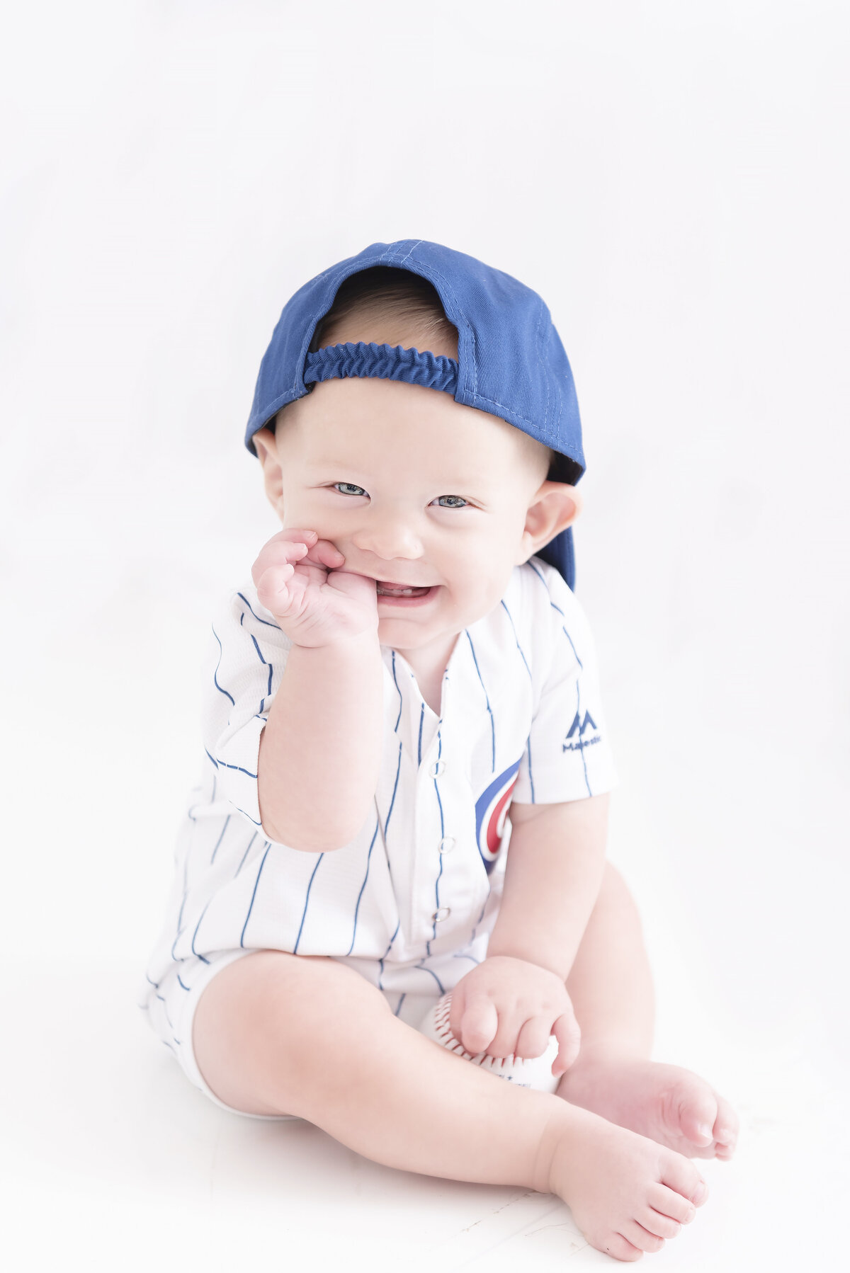 A toddler baby boy sitting in a baseball uniform and hat in a studio