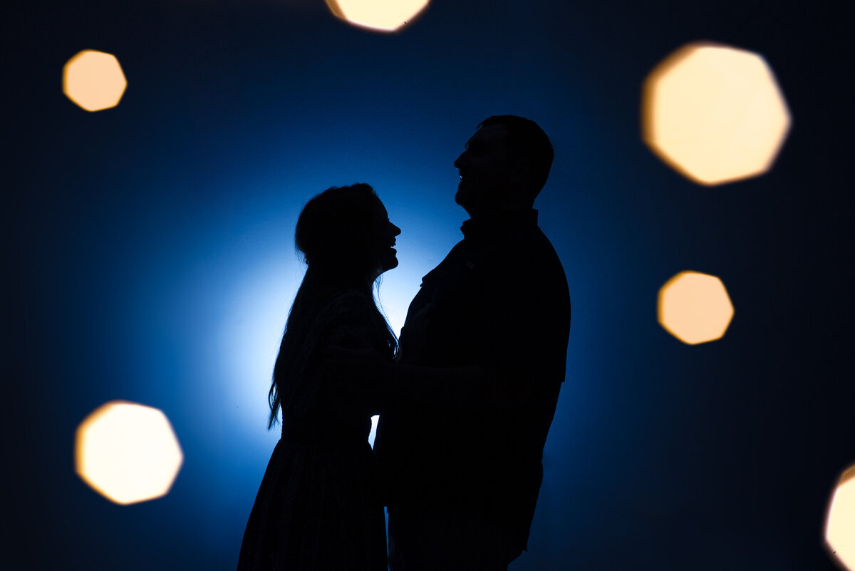 Silhouette-of-an-engaged-couple-laughing-in-front-of-a-blue-lit-background-with-twinkle-lights-in-the-foreground