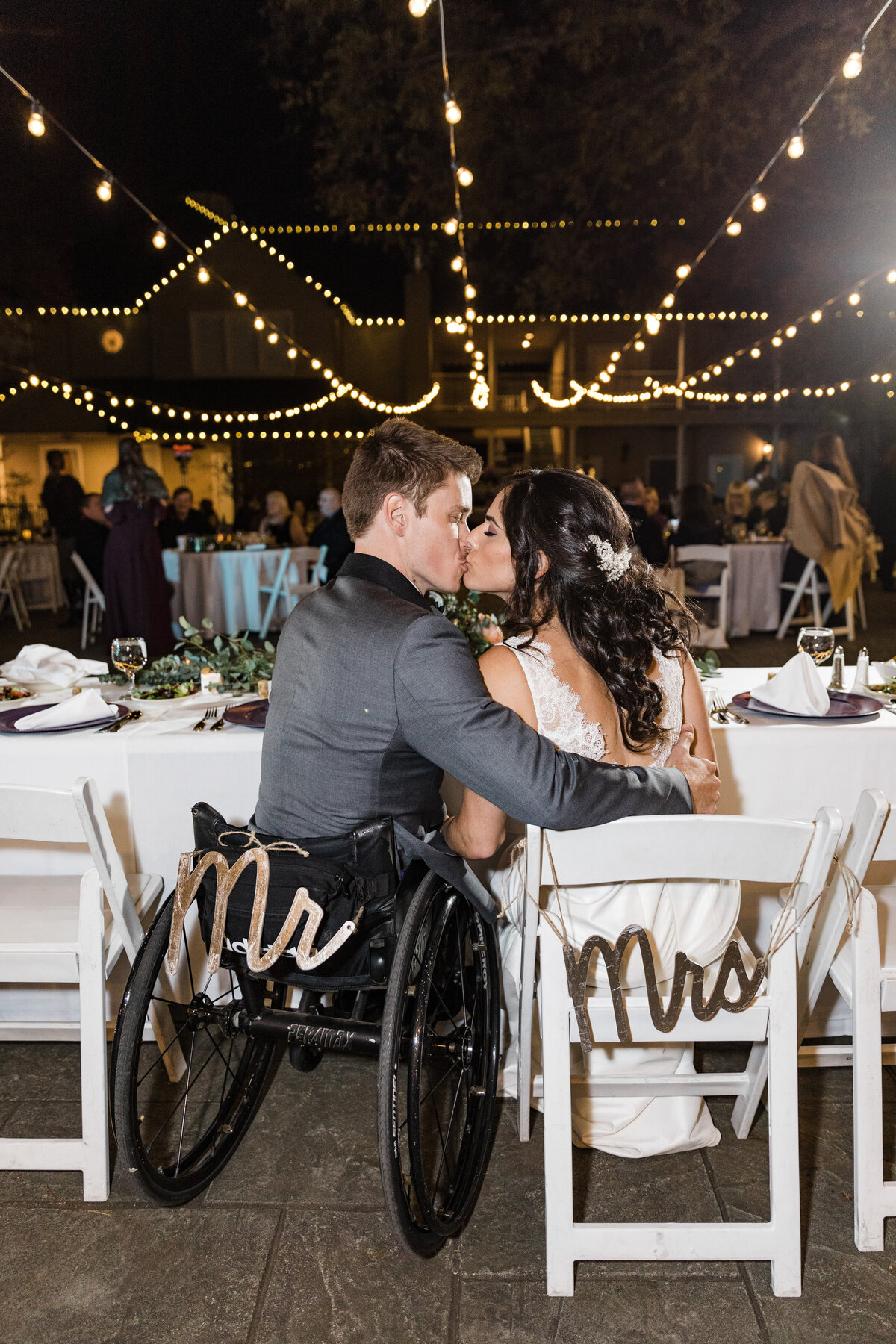 A portrait of a bride and groom sharing a kiss at their head table during their wedding reception in Dallas, Texas. The bride is on the right and is wearing a intricate white dress and a hairpiece. The groom is on the left and is wearing a grey suit. The bride is seated in a white chair with a "Mrs." sign hanging off the back while the groom is seated in his wheelchair with a "Mr." sign hanging off the back. In the background, many strands of cafe lights crisscross overhead of all the guests and their reception tables.