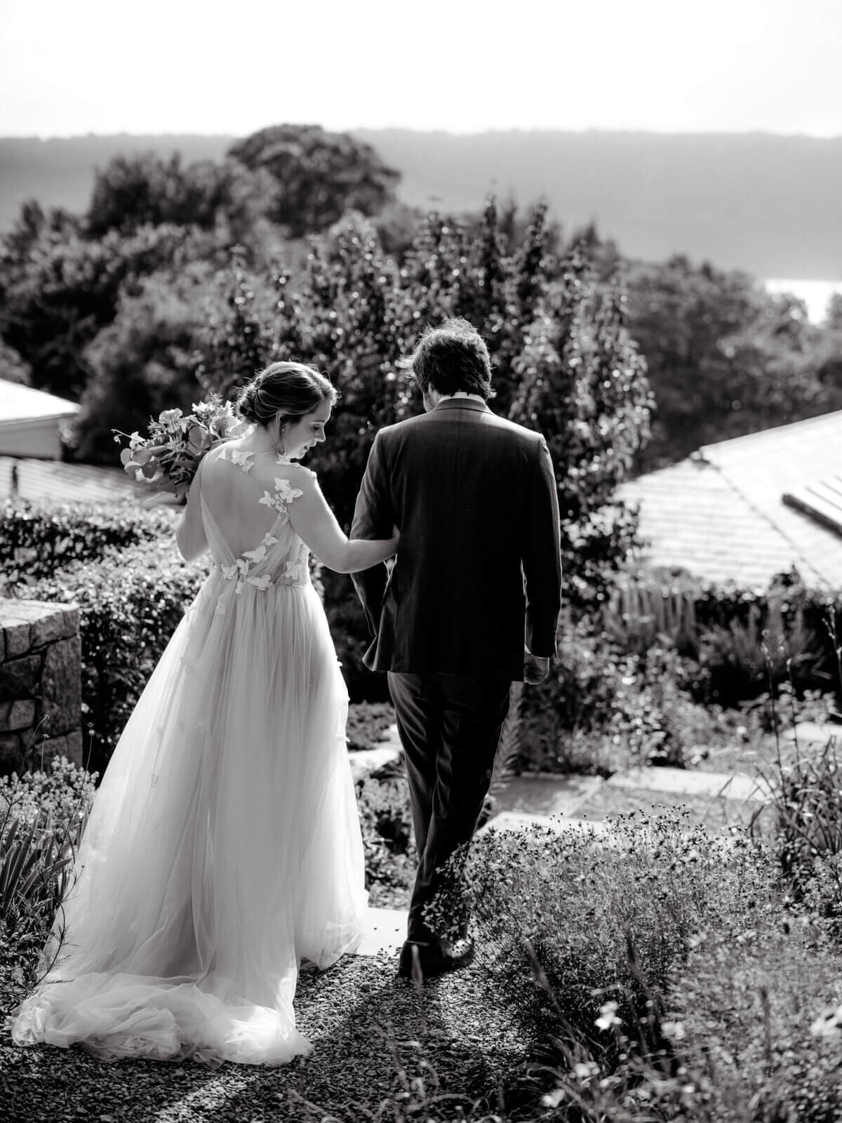Back view of the bride and the groom, while the bride, looking down sideways, is holding on the groom's arms, at a garden