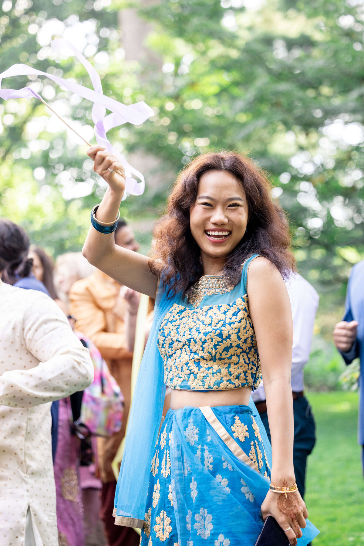 A woman in a blue and gold traditional outfit joyfully waves a white ribbon at an outdoor event. She is smiling widely, surrounded by greenery and other people in colorful attire.