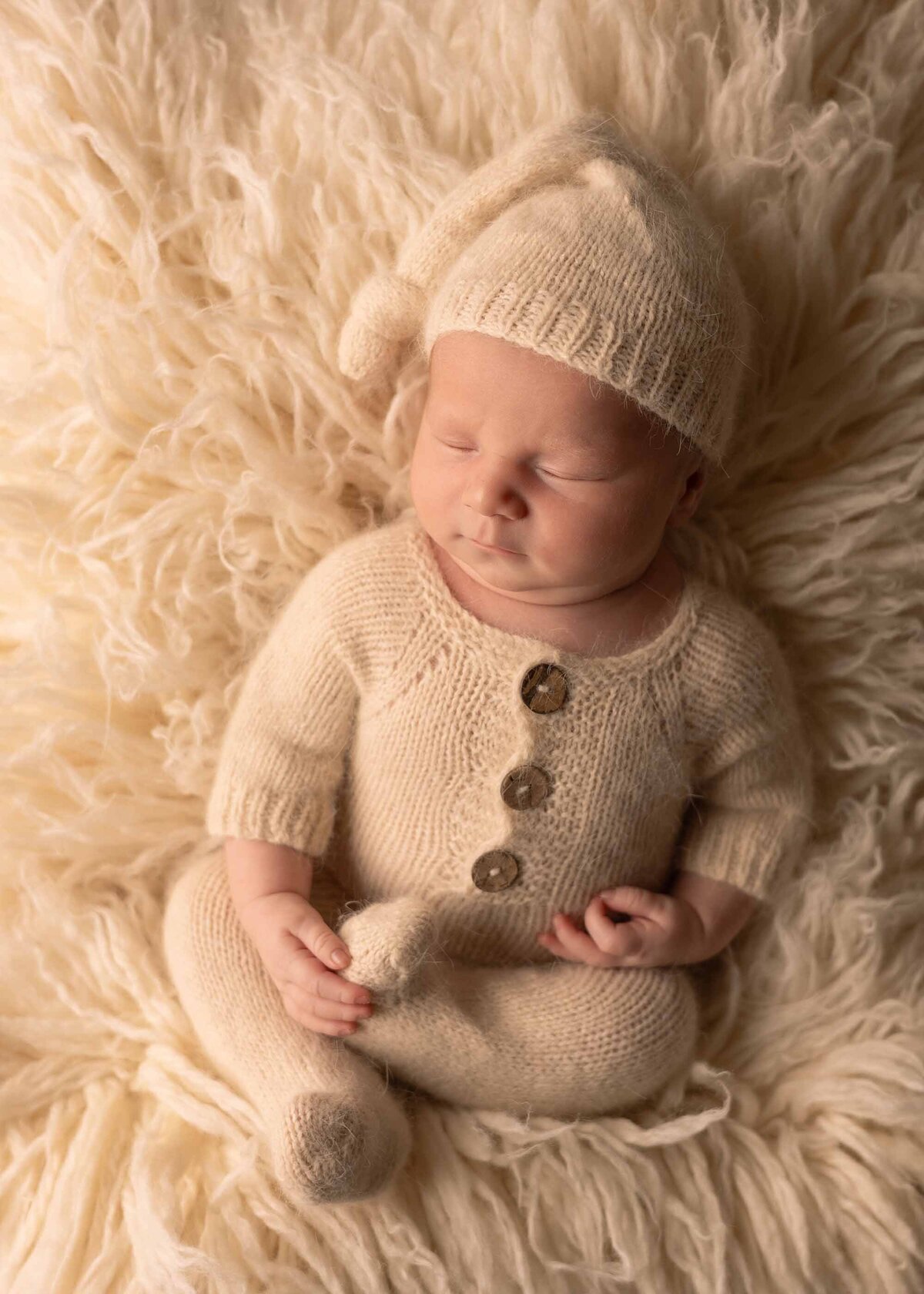 Maddie Rae Photography baby sleeping. he is wearing a neutral outfit and is laying on a neutral colored fluffy rug. his legs are crossed and he is holding one foot. he has a little matching hat on as well