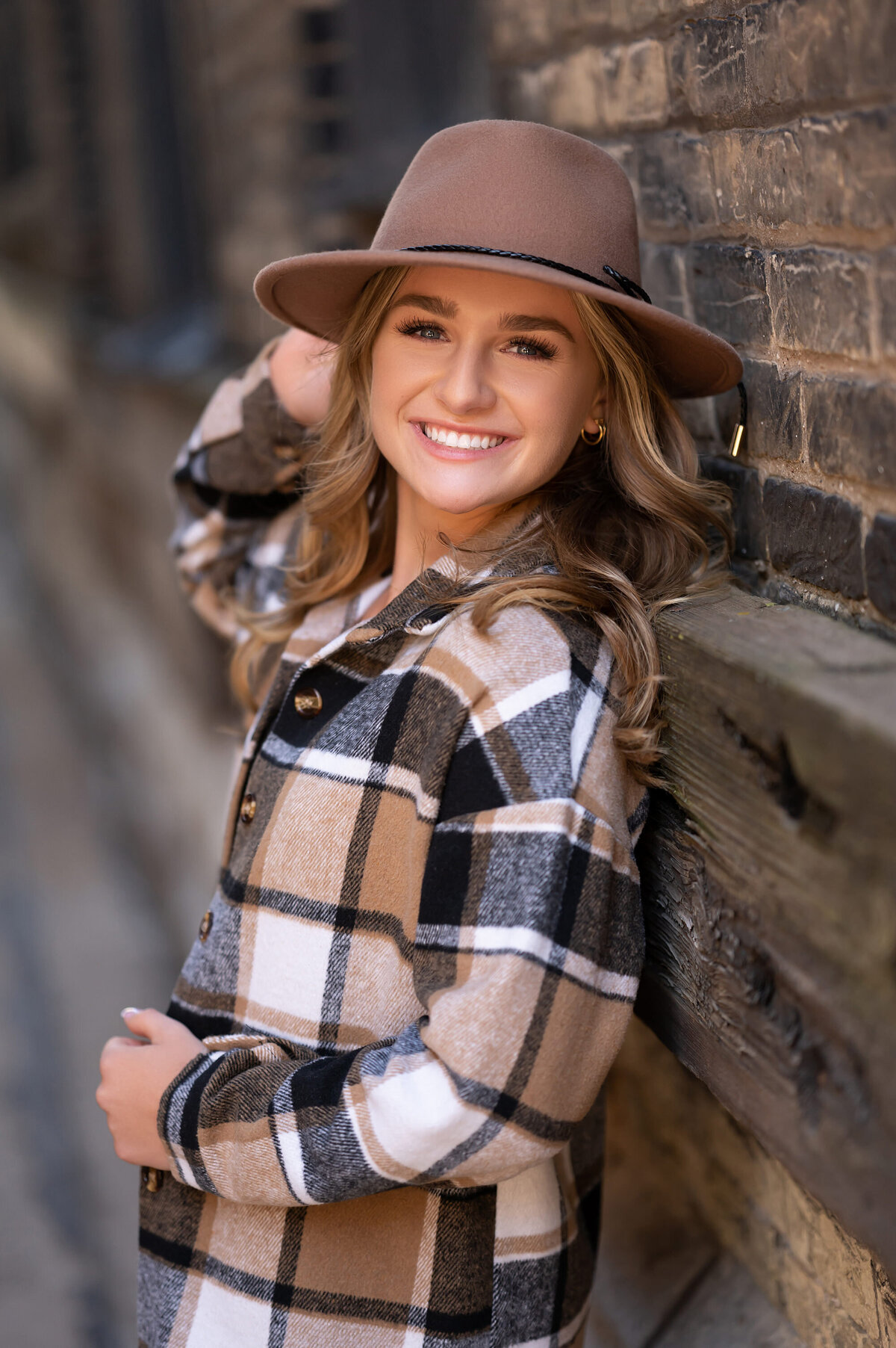 A young woman from Waukesha West High School leans against a brick wall in Milwaukee's Third Ward wearing neutral flannel for her senior portraits.
