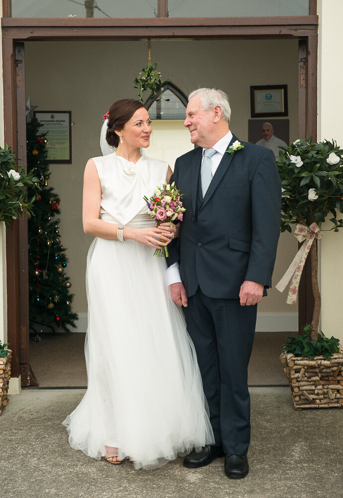 brunette bride wearing an a-line, satin and tulle wedding dress standing at the church door with her dad who is wearing a dark nay suit