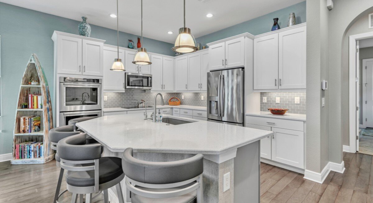 The interior of a kitchen with white cabinets, light teal walls, and stainless steel appliances.  All the lights are on and the room is open and bright.