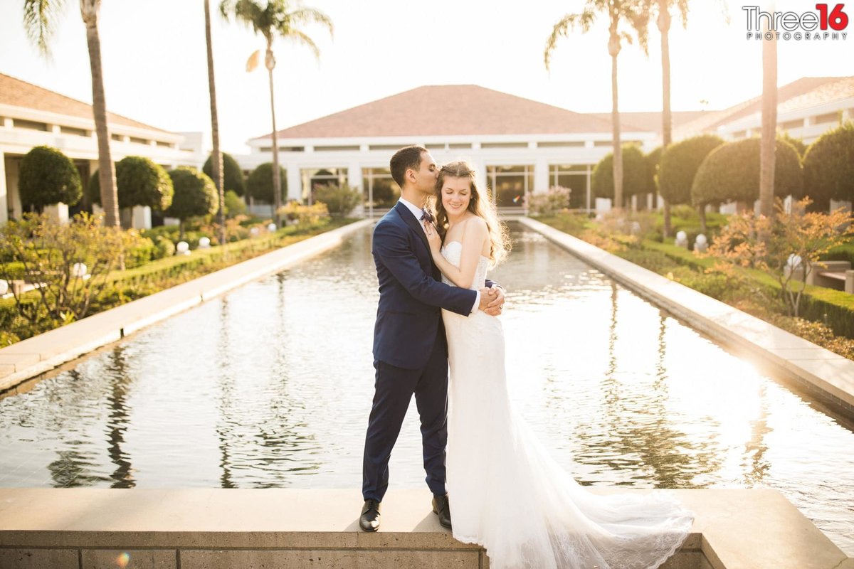 Groom embraces his Bride while standing on the wall surrounding the reflection pool at the Richard Nixon Library