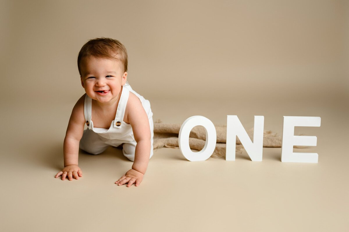 1-year-old Milestone session were a little boy is dressed and white overalls crawling towards the camera with the o n e letters beside him on a brown Studio background