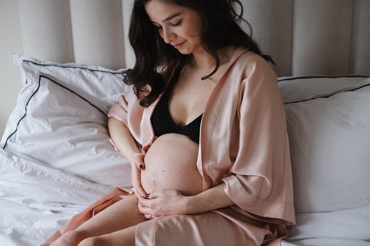 Woman wearing pink silk robe sits in bed looking down at her pregnant belly