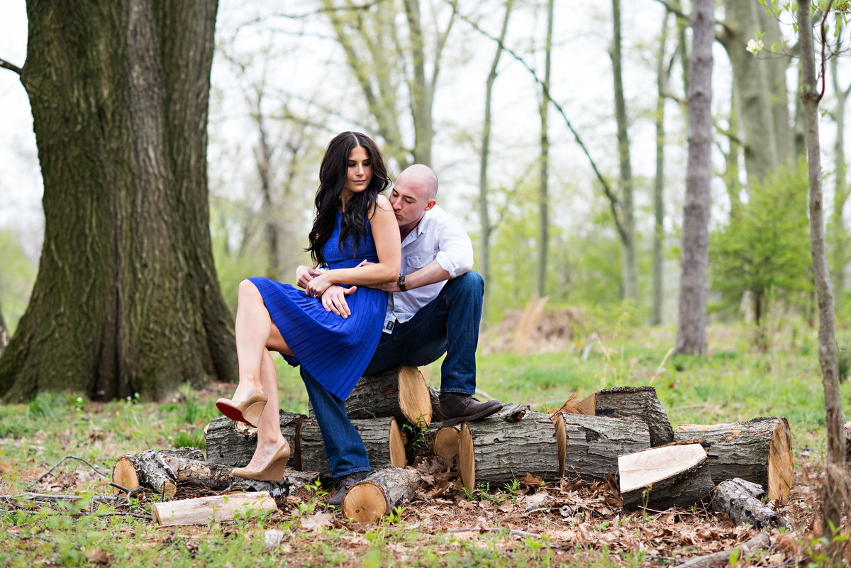 A bride to be sits on her fiance's lap on top of chopped logs in the woods.
