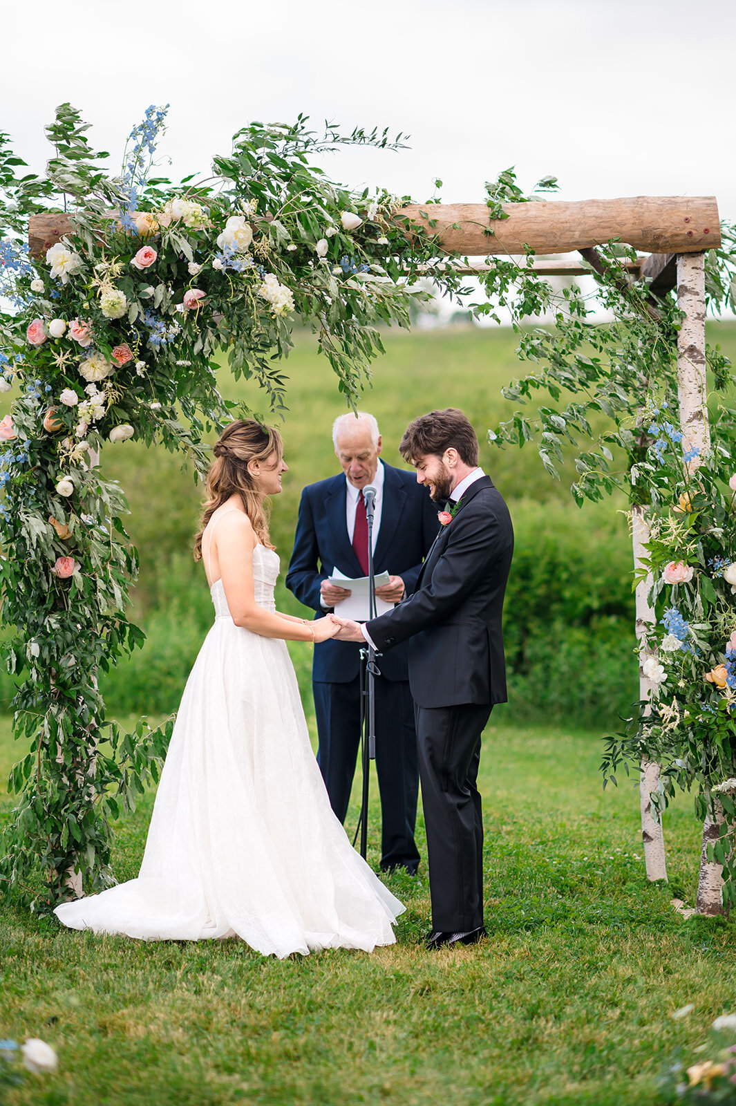 A bride and groom exchanging vows under a rustic wooden arch decorated with flowers.