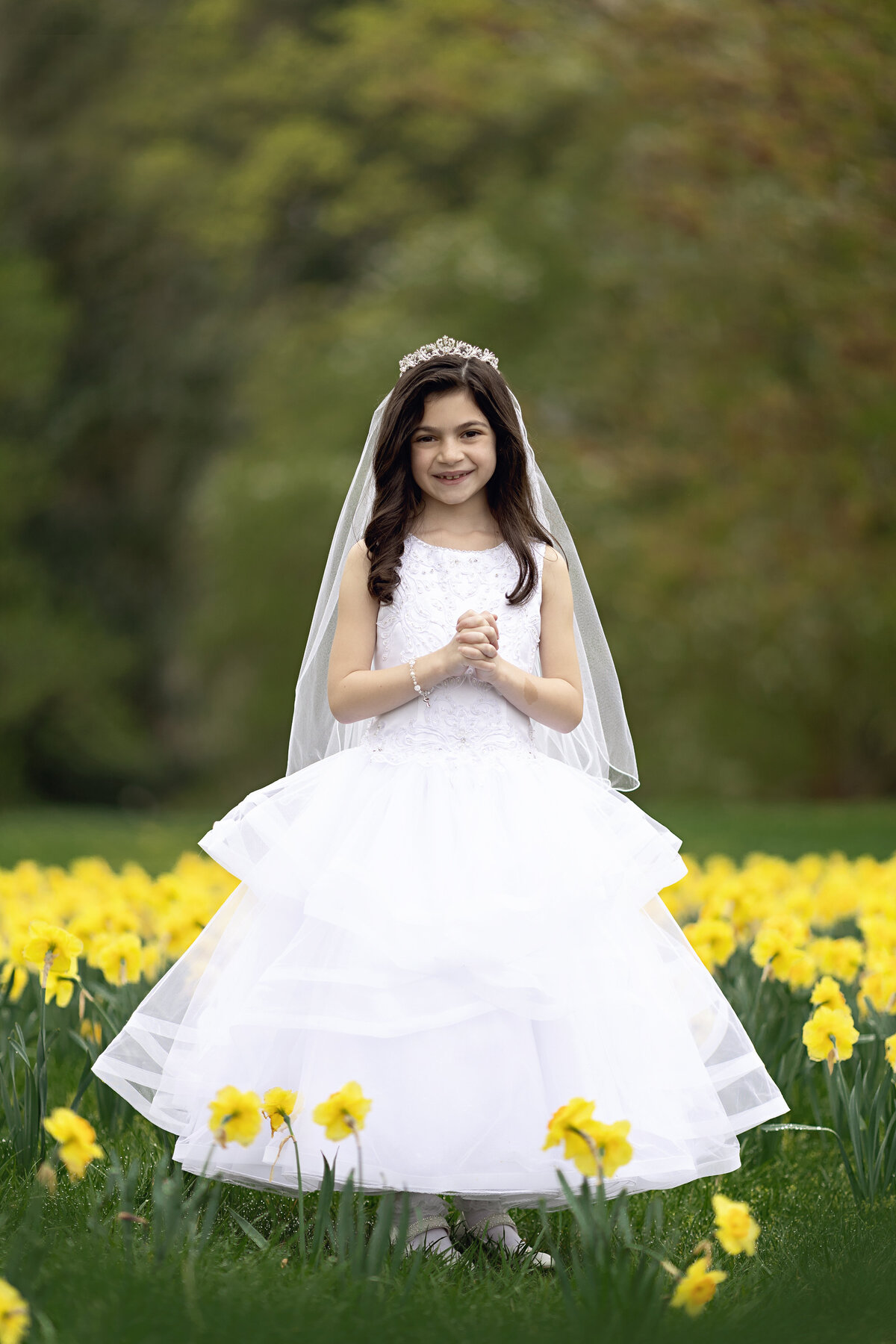 A young dark haired girl stands in a field of yellow wildflowers in a communion dress tiara and veil