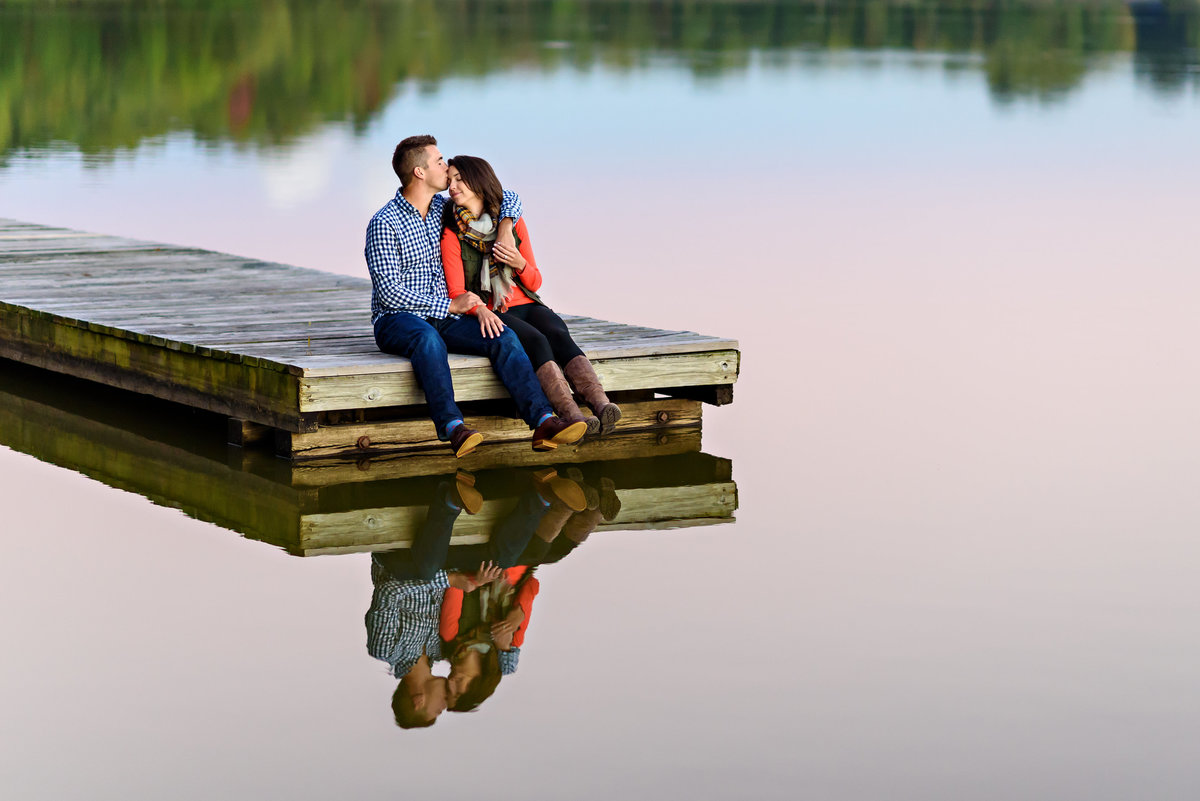 A loving couple sit on the edge of dock in a lake and kiss.