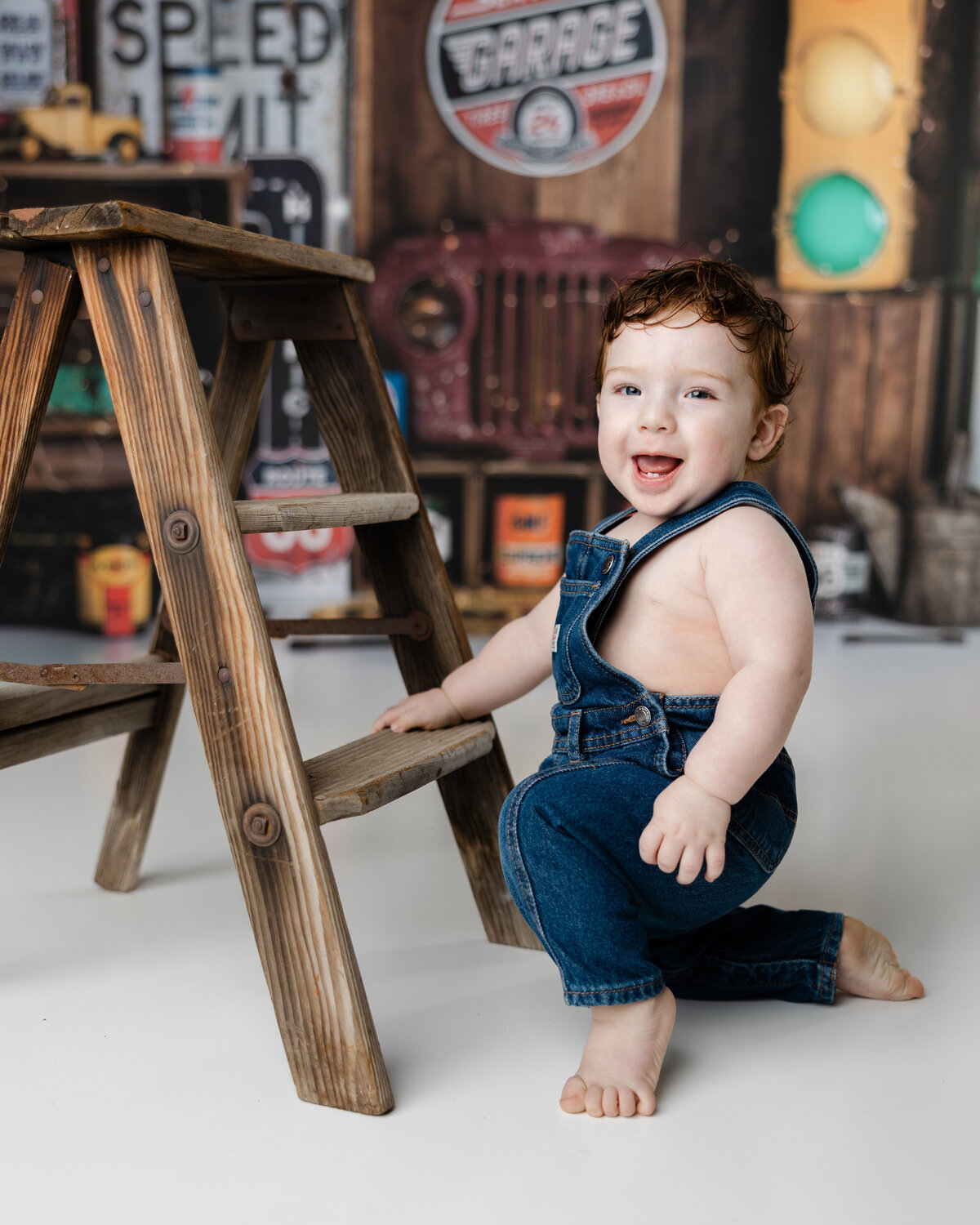Baby boy kneeling and laughing beside a wooden step ladder, dressed in blue overalls with a rustic garage-themed backdrop. Photograph by Yucaipa cake smash photographer Kaitlyn Dawn Photography.