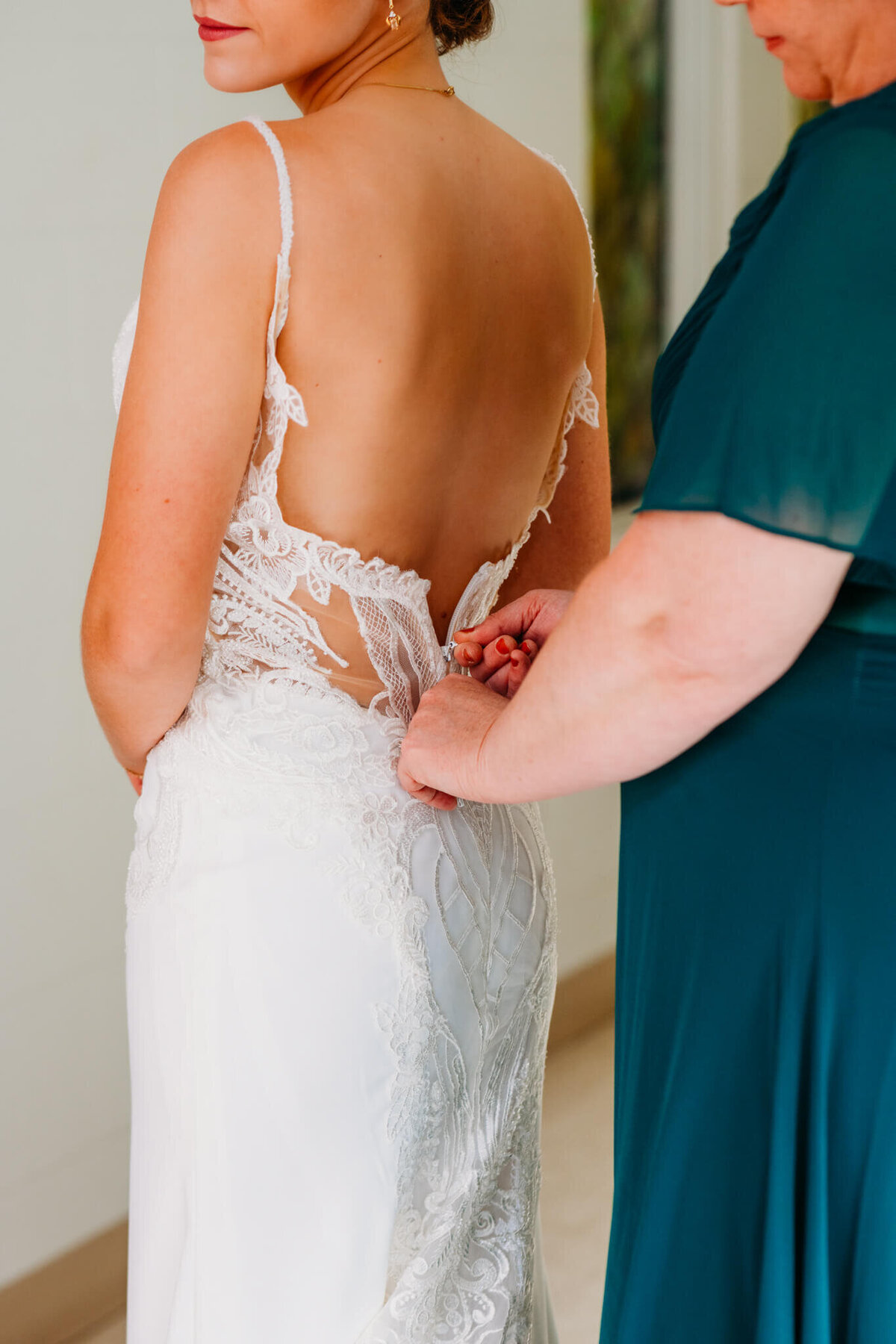 Photo of a brides wedding dress being zipped up by her mom who is wearing a teal dress