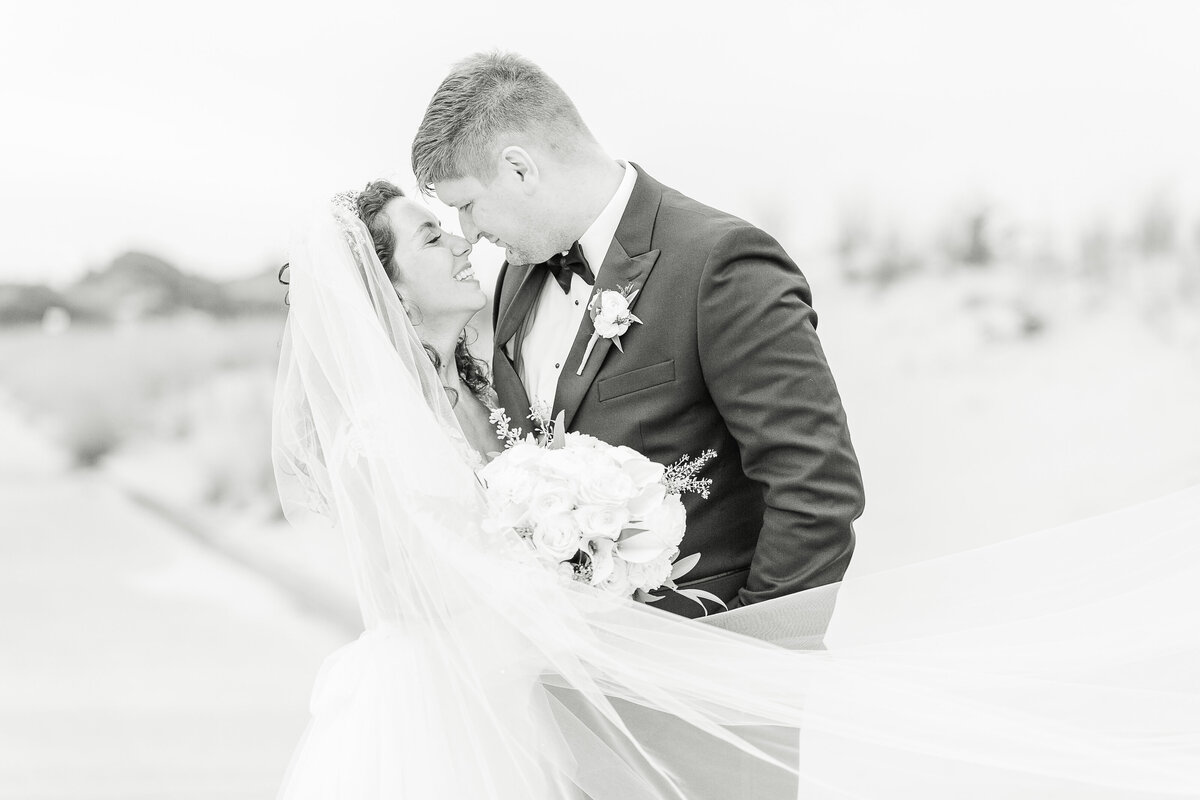 Black and white photo. Bride and groom are captured on the sandy shores of their Dunes Club wedding. Their noses are touching and eyes closed, about to kiss. The bride's veil is blowing in front of them. Captured by best Rhode Island wedding photographer Lia Rose Weddings.