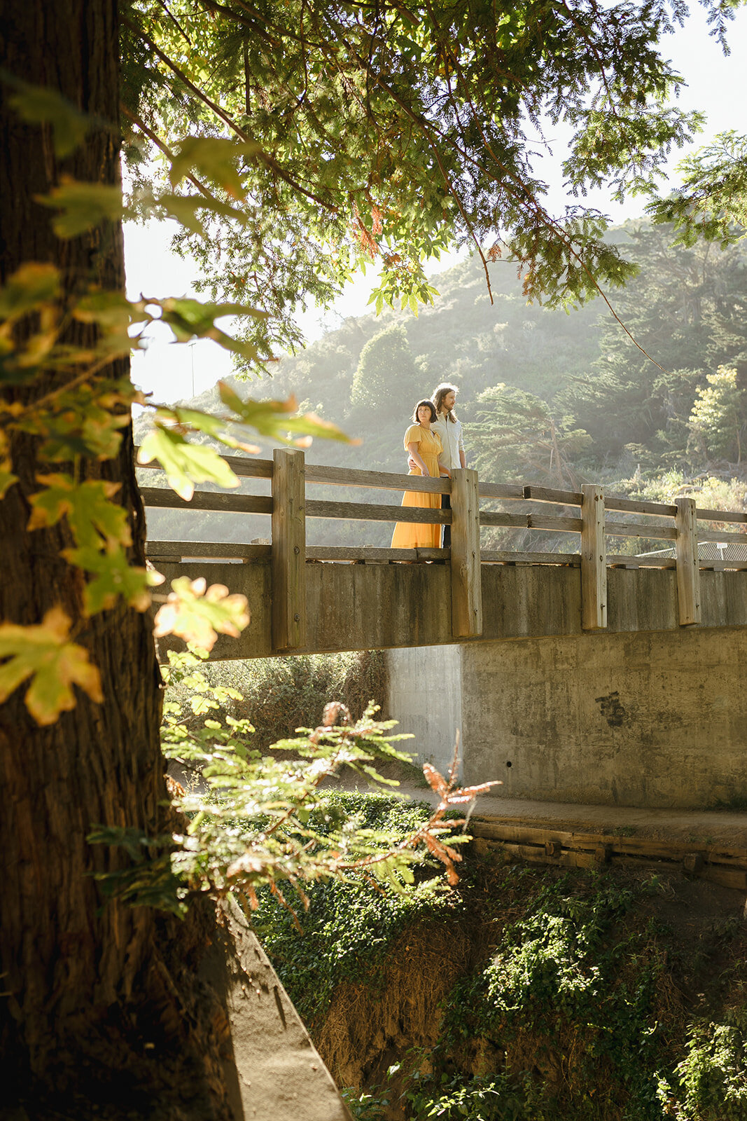 Woman in yellow dress stands next to partner on a bridge, backlit by the sun, in Big Sur, California.