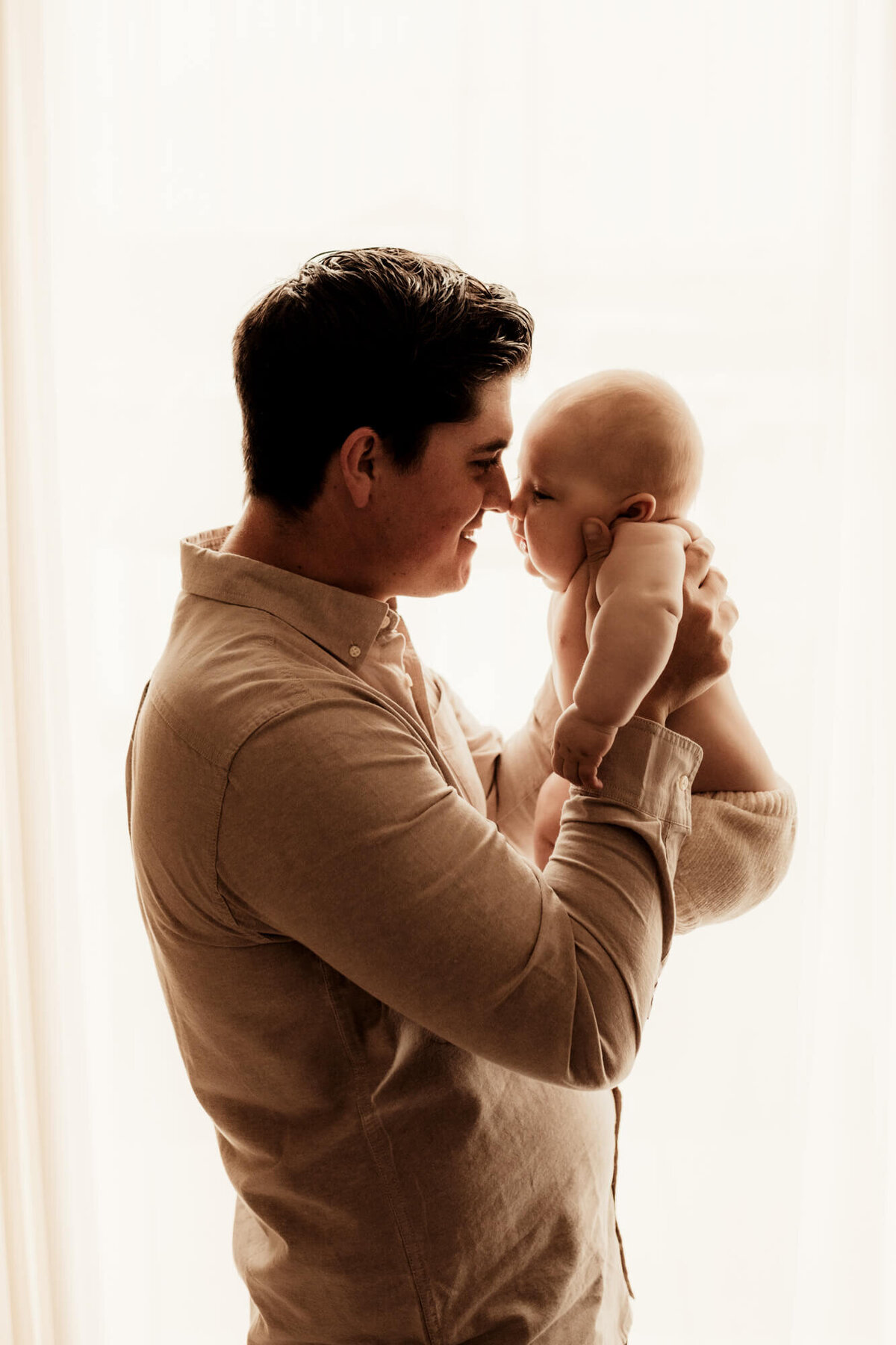 Father and baby touch noses next to a window.