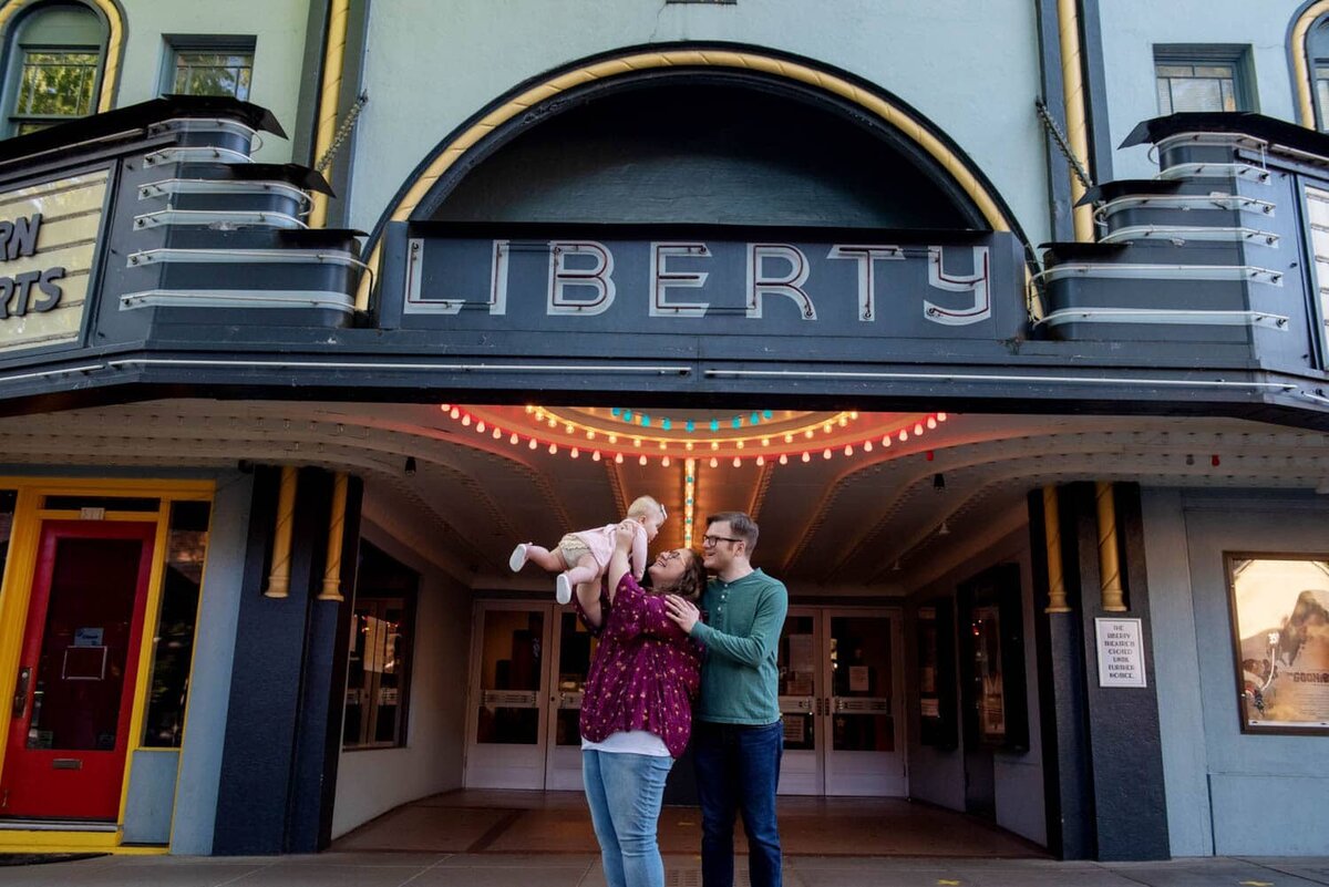 Mom tossing baby in air with dad behind her, smiling in front of Liberty Hall in Downtown Camas.