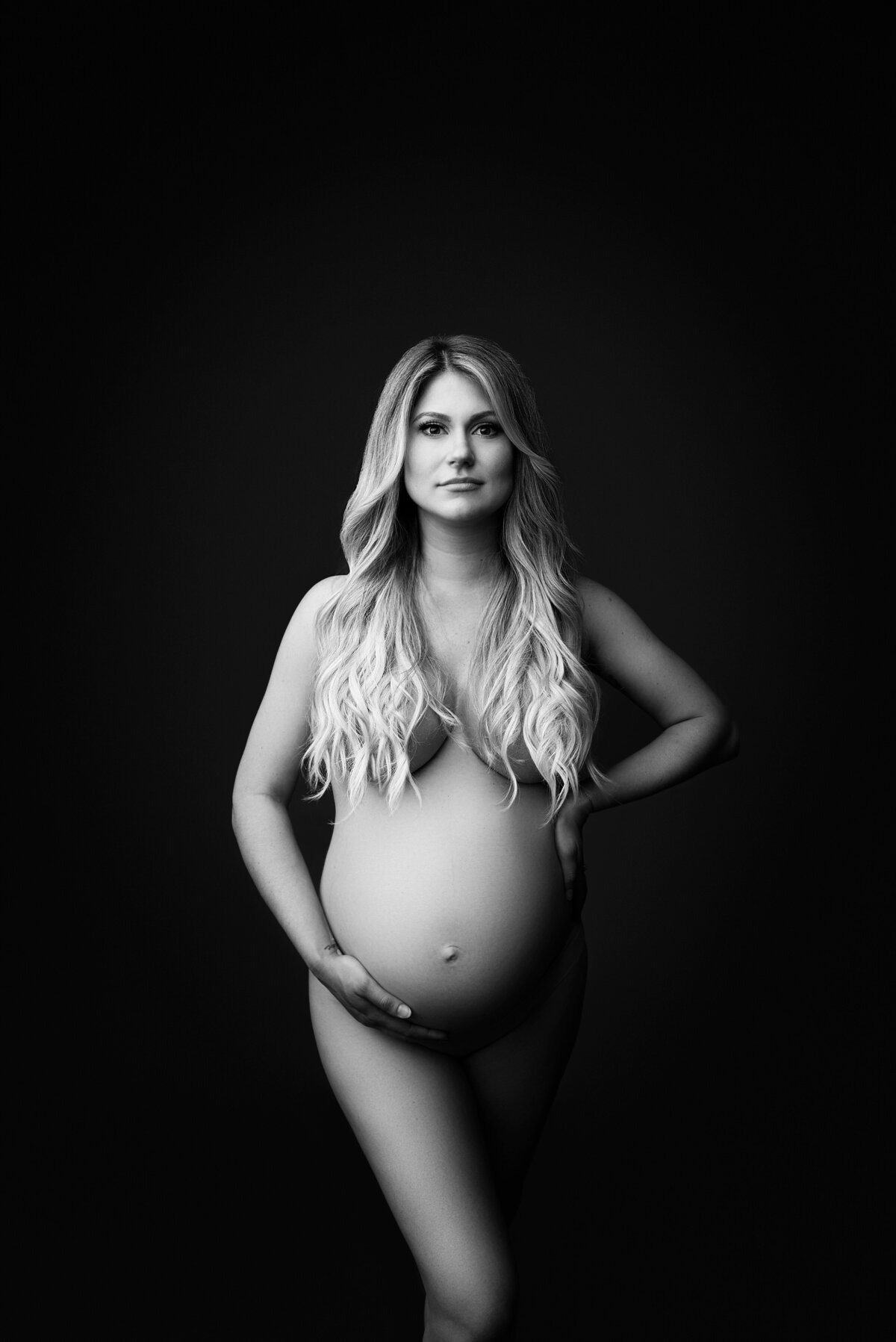 New Jersey's best maternity photographer Katie Marshall captures expectant mom for fine art maternity photos. Mom is standing facing the camera bare. Her long blonde wavy hair is pushed to the front to hide her breasts and her front knee extended to hide her intimate parts. One hand is resting underneath her bump, the other on the small of her back. The left half of her body is enveloped in light, the right half shadowed to create interest and dimension.