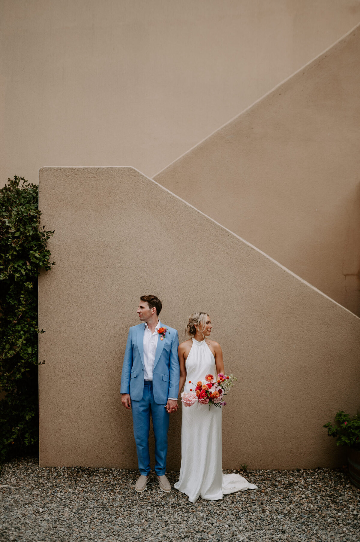 A bride and groom face away from each other next to a Wes Anderson looking set of stairs at Chateau Canet. The groom is wearing a blue suit and no tie where the brides dress is sleeveless as she is also carrying pink and orange flowers.