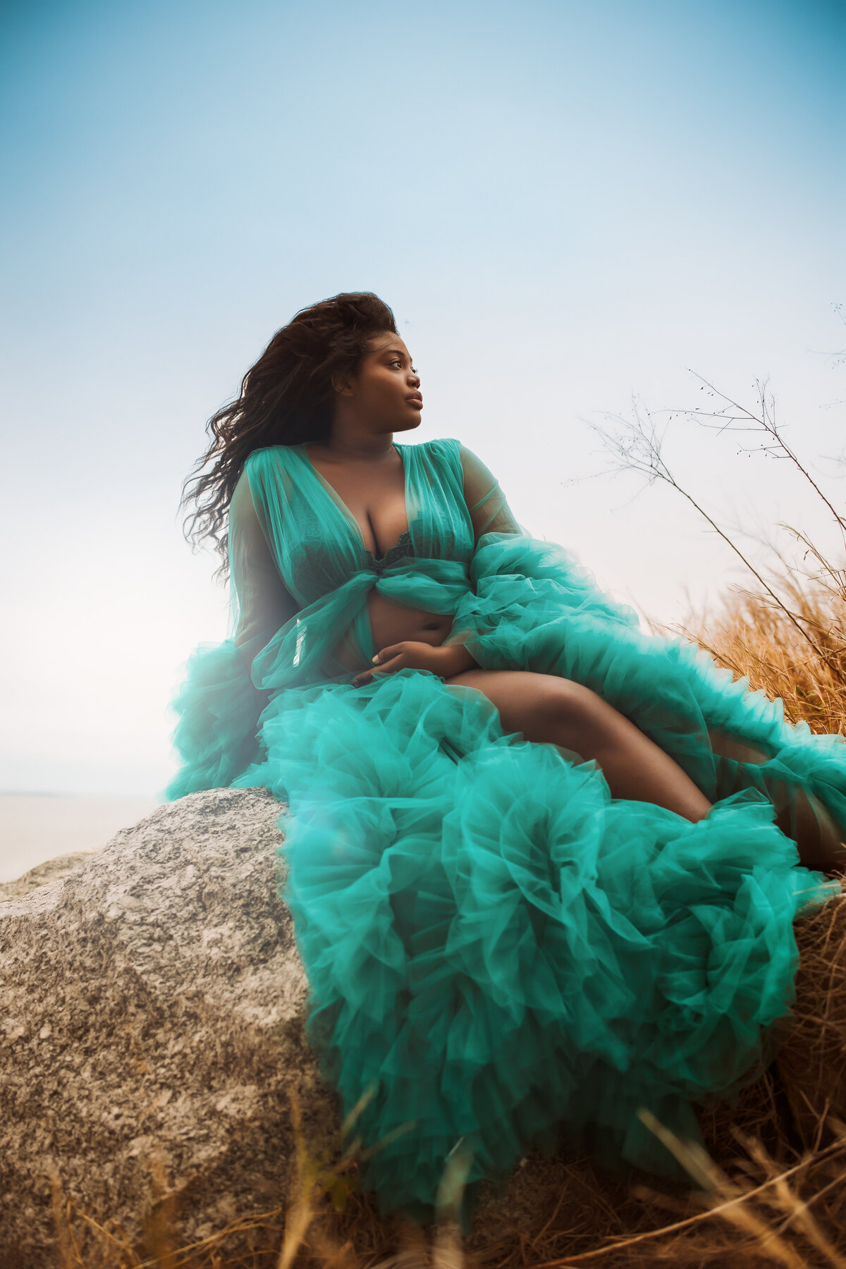 stunning expecting mother wearing a teal tulle robe sitting on a  rock at fred howard park for a pregnancy shoot at sunset