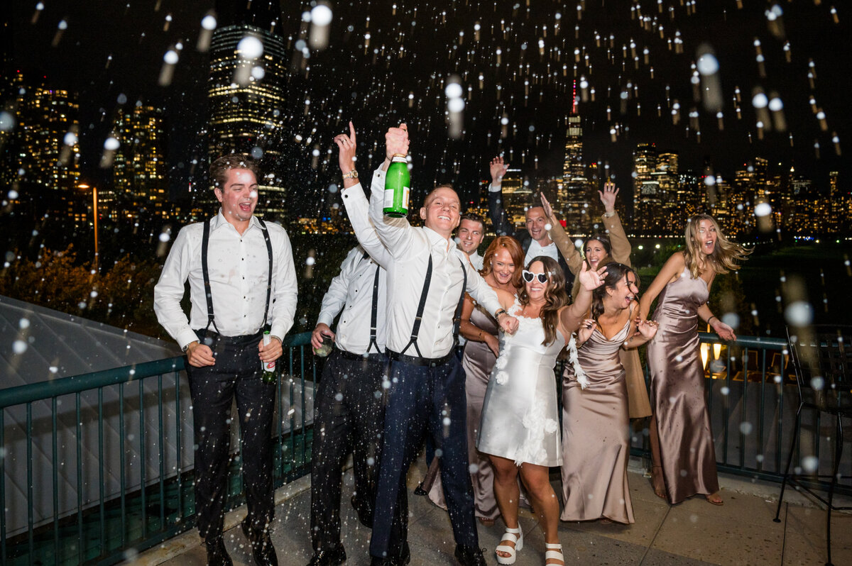 Bride and groom spraying champagne on the balcony of liberty house in jersey city at night with the nyc skyline behind them